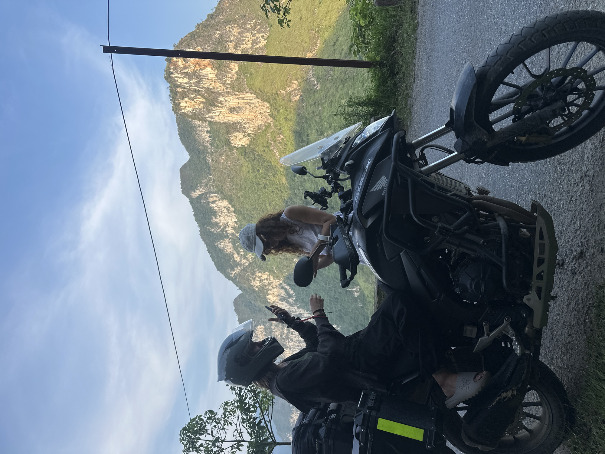 Two individuals are positioned near a parked motorcycle on a paved road. The backdrop features a mountainous landscape, consistent with the terrain in Đường Thượng Commune, Vietnam. One individual, wearing a helmet, is seated on the motorcycle and appears to be using a mobile phone. The other individual, wearing a cap, is standing next to the motorcycle.