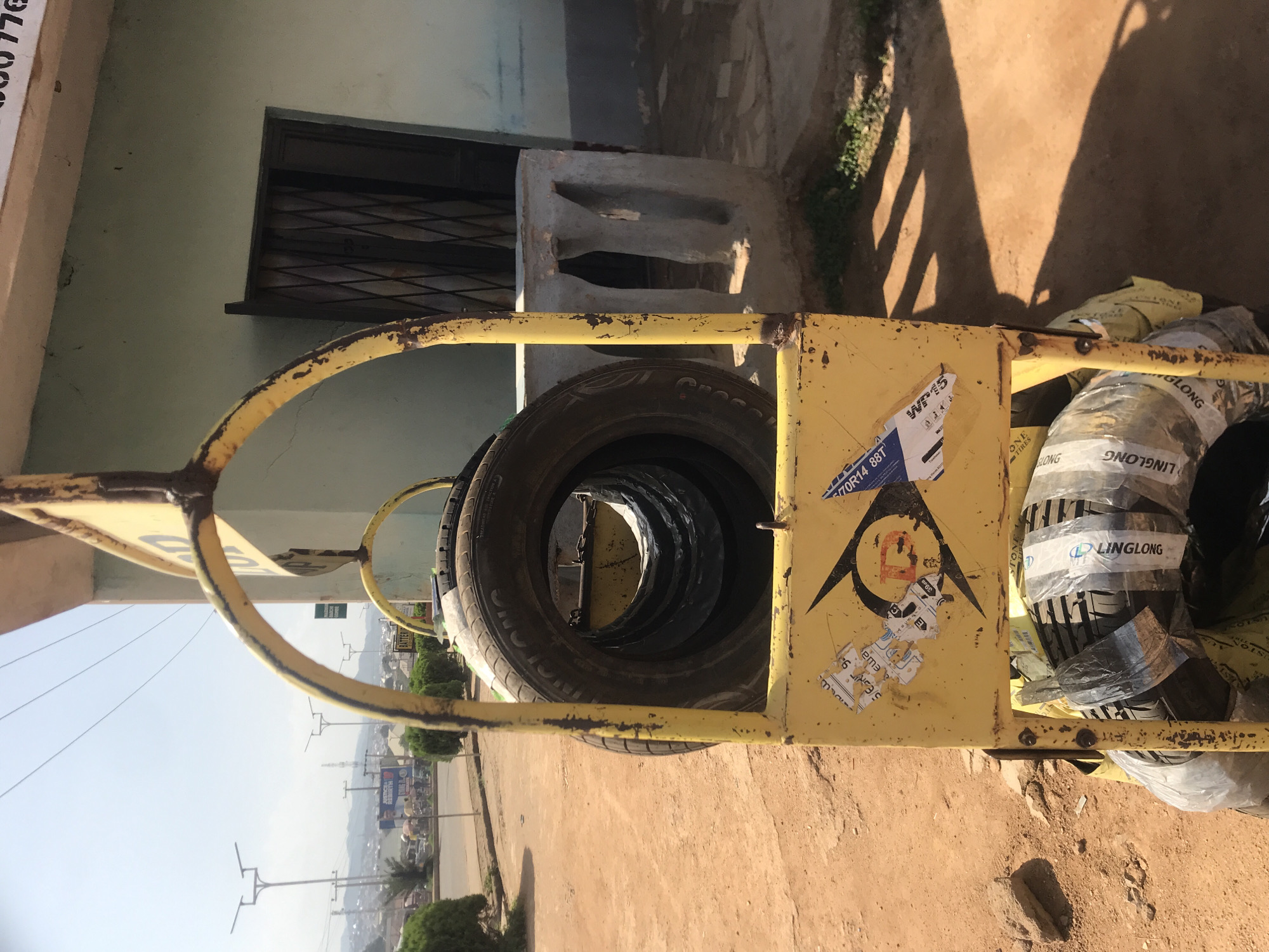The image depicts a tire stand filled with tires, situated outdoors in Rafin Par, Nigeria. The stand is constructed from weathered yellow metal, showing signs of rust and wear. It holds several tires, some stacked within each other, and others wrapped with plastic and tape. Stickers, including one with a circular logo and text, are affixed to the stand.
In the background, a light green building with a closed, barred doorway is visible. To the left of the building, a concrete structure with arched openings stands. The ground is covered in reddish-brown dirt, with scattered stones. Further in the distance, a street with sparse traffic, lampposts, and various signs can be observed, suggesting an urban or semi-urban environment. The sky is clear and bright, indicating daylight. There are no people clearly visible in the immediate vicinity of the tire stand.