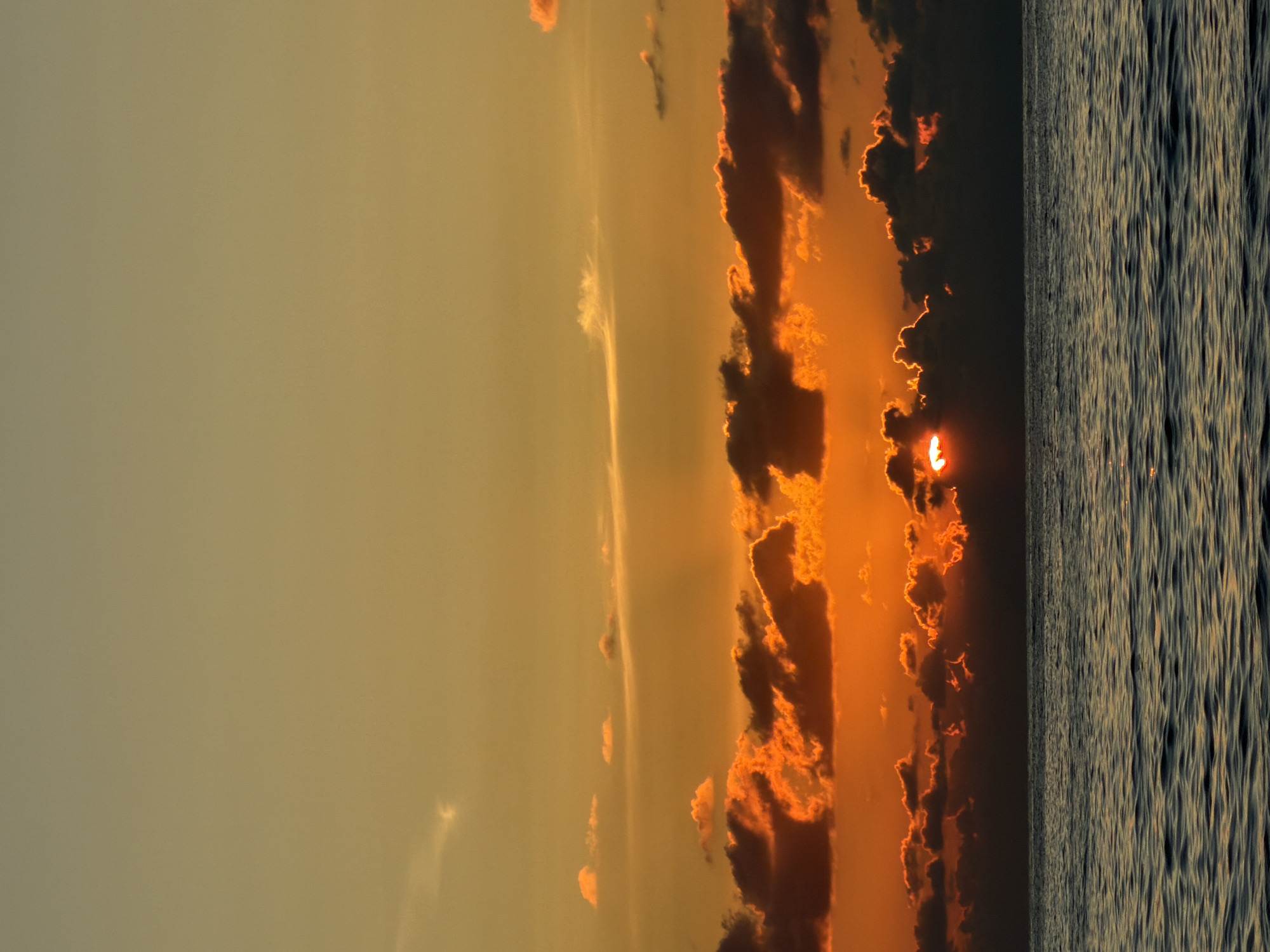 This is a photograph of a sunset over the Black Sea in Sochi, Russia. The sky is a gradient of warm colors, from a soft yellow at the top to a fiery orange and red closer to the horizon. Clouds are scattered across the sky, with some illuminated by the setting sun and others appearing as dark silhouettes. The sun itself is partially obscured by clouds, creating a dramatic glow. The water of the Black Sea is dark and rippling, reflecting the colors of the sky. The horizon line between the sea and the sky is sharp and clear. There are no people or man-made objects visible in the image, making it a natural landscape scene.