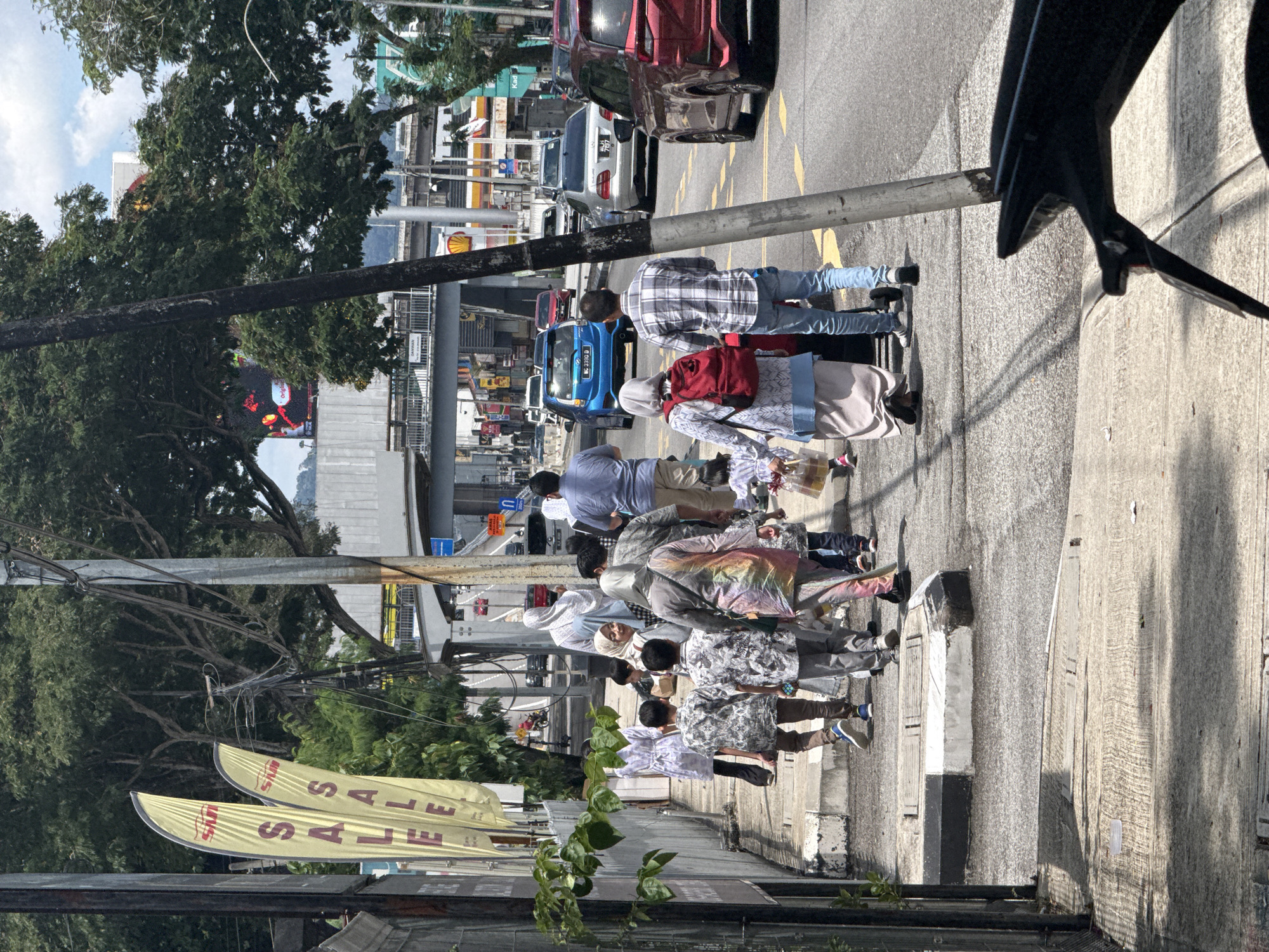 The image shows a street scene in Kuala Lumpur, Malaysia. A group of at least ten people are walking on the sidewalk. Some appear to be families with children. Most people are wearing casual clothing, and several women are wearing headscarves. There are banners with the word "SALE" visible, suggesting a retail environment. Cars are parked along the street, and others are moving in traffic. There is also a Shell gas station visible. The weather is bright and sunny. Buildings and trees line the street. Overhead power lines and poles are also visible.