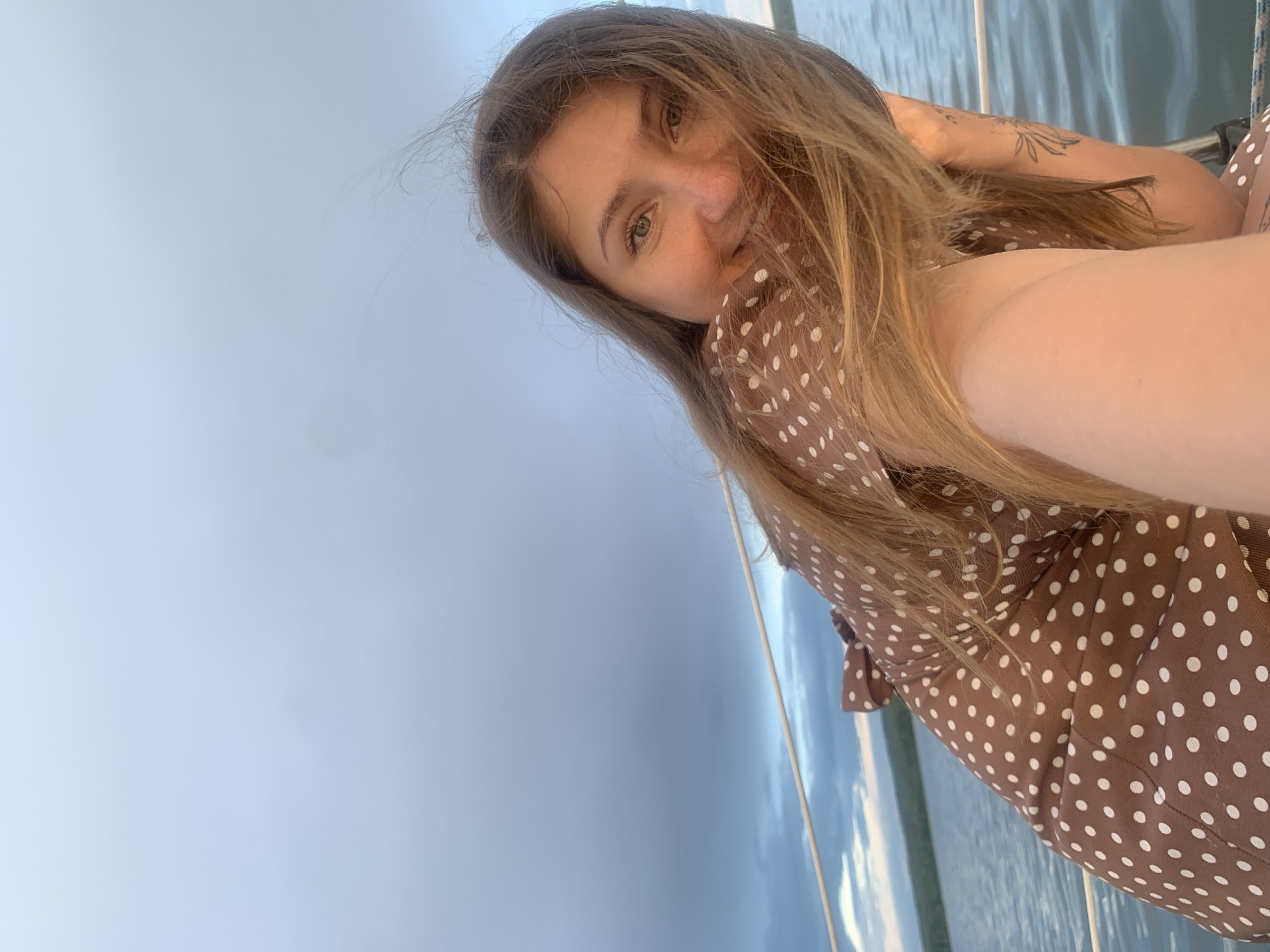 The image shows a young woman on a boat in Chaykovsky, Russia. The woman has long blonde hair and is wearing a brown polka dot dress. She is smiling and looking at the camera. The boat is on a body of water, and the sky is cloudy. In the background, there is a shoreline with trees.