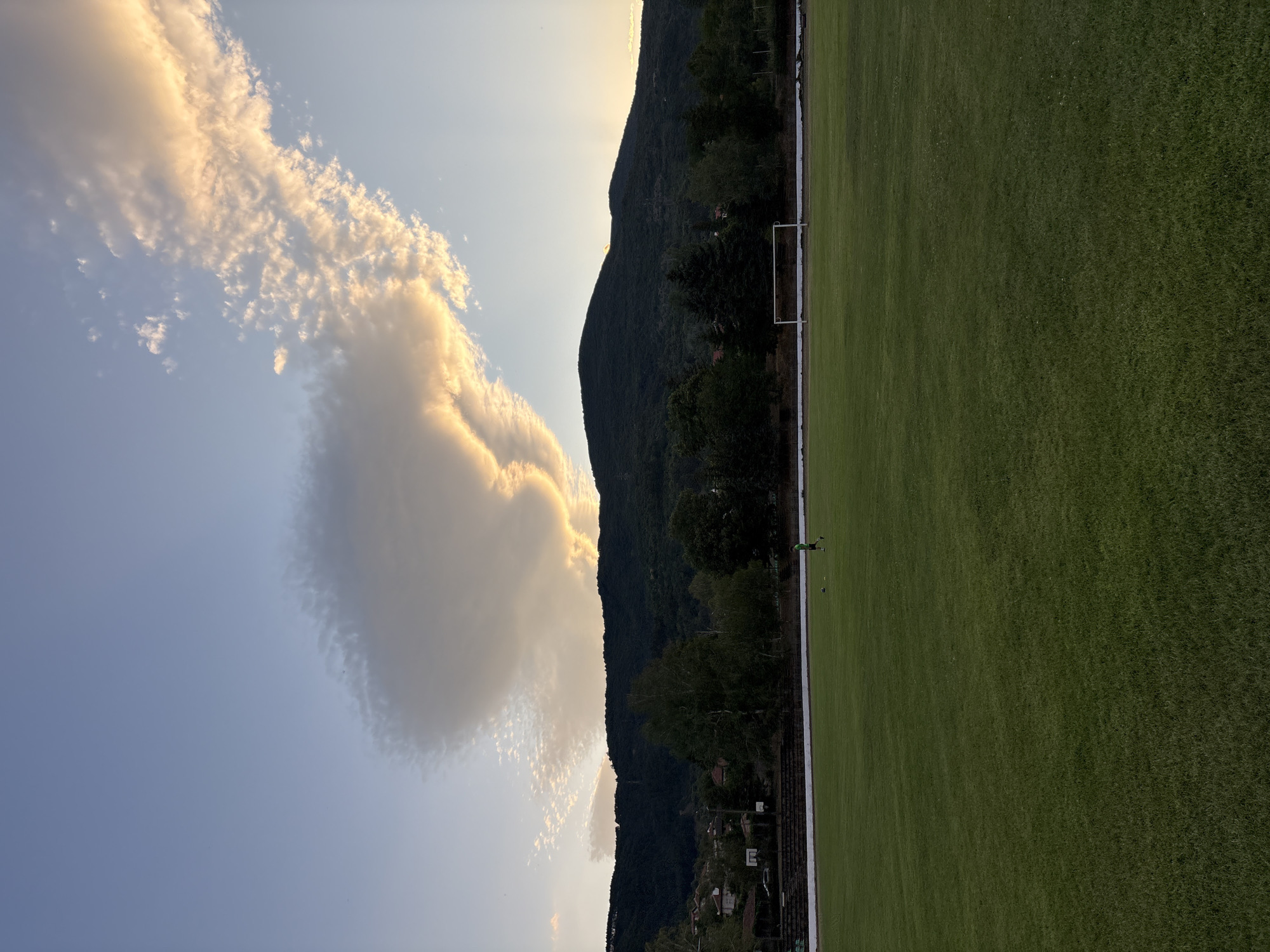 This image depicts a sports field in Boboshevo, Bulgaria, under a sky adorned with dramatic clouds. In the foreground, a lush green grass field stretches out, indicating it is likely a soccer field, evidenced by a goalpost visible in the mid-ground. A single individual, dressed in a green shirt and dark shorts, stands on the field, appearing to be a player. The mid-ground also features a line of bleachers or seating areas, partially obscured by trees and the structures of the town. In the background, a dense, tree-covered mountain range dominates the horizon. The sky is a prominent feature, with a large, luminous cloud formation catching the late afternoon or early evening light, casting a warm glow and creating a striking visual contrast against the deeper blues and grays of the sky. The overall atmosphere is serene, with the natural landscape and the athletic setting converging.