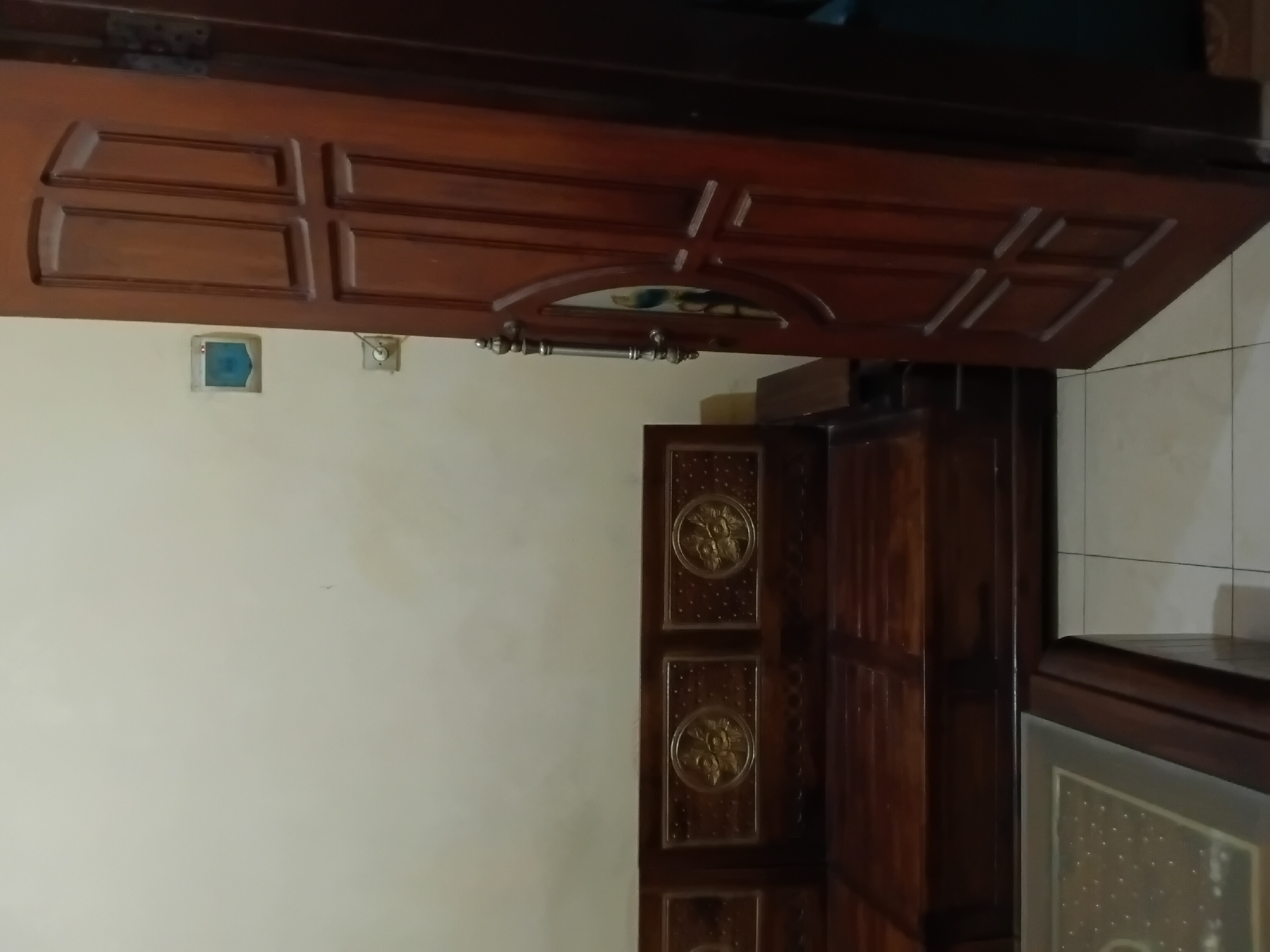 The image depicts an interior scene in a room, likely a living room or common area. The main subjects are a dark wooden sofa with ornate circular carvings on its backrest, and a wooden door which is slightly ajar on the right side of the frame. The door has a traditional design with multiple raised panels and a metallic handle with decorative elements. On the wall to the left of the door, there is a light-colored electrical outlet and a square-shaped device with a blueish square in the center. The floor is tiled with light-colored square tiles. A portion of a wooden coffee table with a glass top is visible in the lower left corner. The walls are a neutral, light color. The lighting suggests an indoor environment with artificial light or ambient daylight filtering in. The scene is static, with no people or apparent activity. This scene is located in the city of Trenggalek, Indonesia.