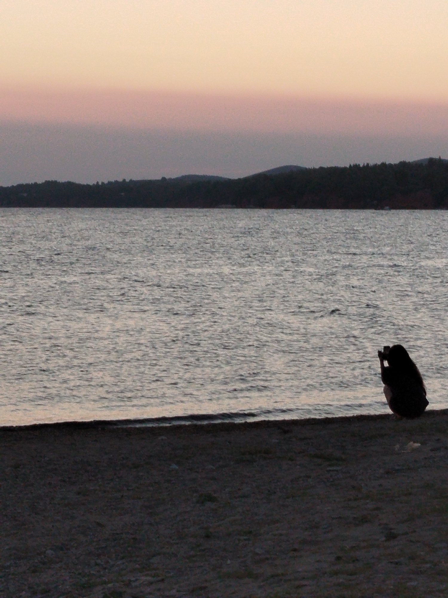 The image shows a silhouetted person sitting on a beach, facing a body of water. The person is holding a camera and appears to be taking a photo. The water is calm and reflects the pale pink and orange hues of the sky. In the distance, a dark, tree-lined shore is visible. The beach itself is covered in dark sand and pebbles. The scene suggests a peaceful moment at sunset in Sithonia Municipal Unit, Greece.