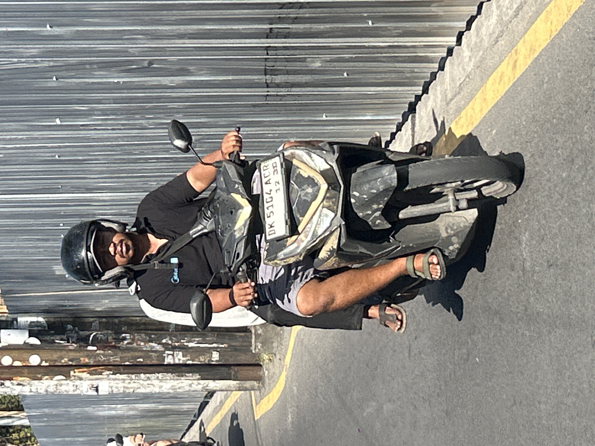 A man wearing a black t-shirt and shorts, and a black helmet, rides a scooter. He is on a road in North Kuta, Indonesia, with a tall metal fence behind him and a yellow line marking the side of the road. The scooter has a license plate that reads "DK 5164 ACR 12 30". The man is holding the handlebars of the scooter, and his feet are on the footrests. He is looking forward, and his face is partially obscured by the sun.