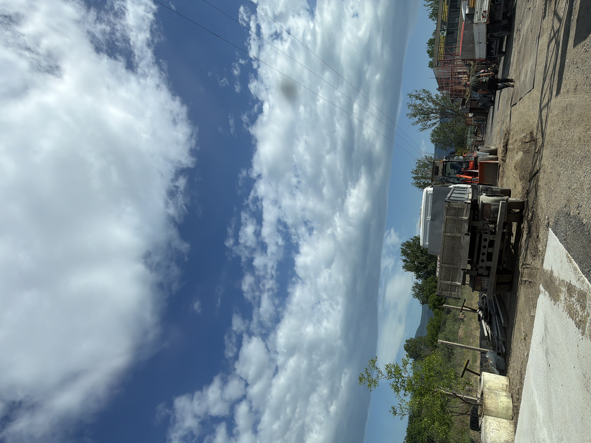 The image depicts a scene in Zlati Voyvoda, Bulgaria. A clear blue sky with scattered white clouds occupies a significant portion of the upper-left area. Several power lines are strung across the upper portion. On the right side, there's an industrial yard with various vehicles, including trucks and a tractor. An individual is standing beside some vehicles. A mix of trees and vegetation is also present. The ground is mostly dirt and gravel, with some concrete slabs.