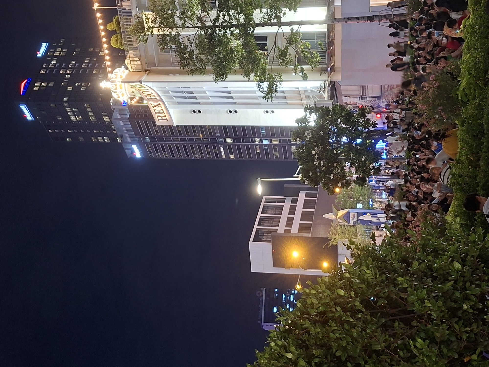 This is a nighttime outdoor scene in Thủ Đức City, Vietnam, featuring a crowd of people gathered in front of the Rex Hotel. The Rex Hotel is illuminated, with its name prominently displayed. Adjacent to it is a tall building with the "Keppel" logo illuminated at the top. The sky is dark, indicating it is night. The foreground is occupied by lush green bushes and trees. The crowd appears to be looking towards something, possibly a performance or a public event. The lighting from the buildings and streetlights creates a vibrant atmosphere.