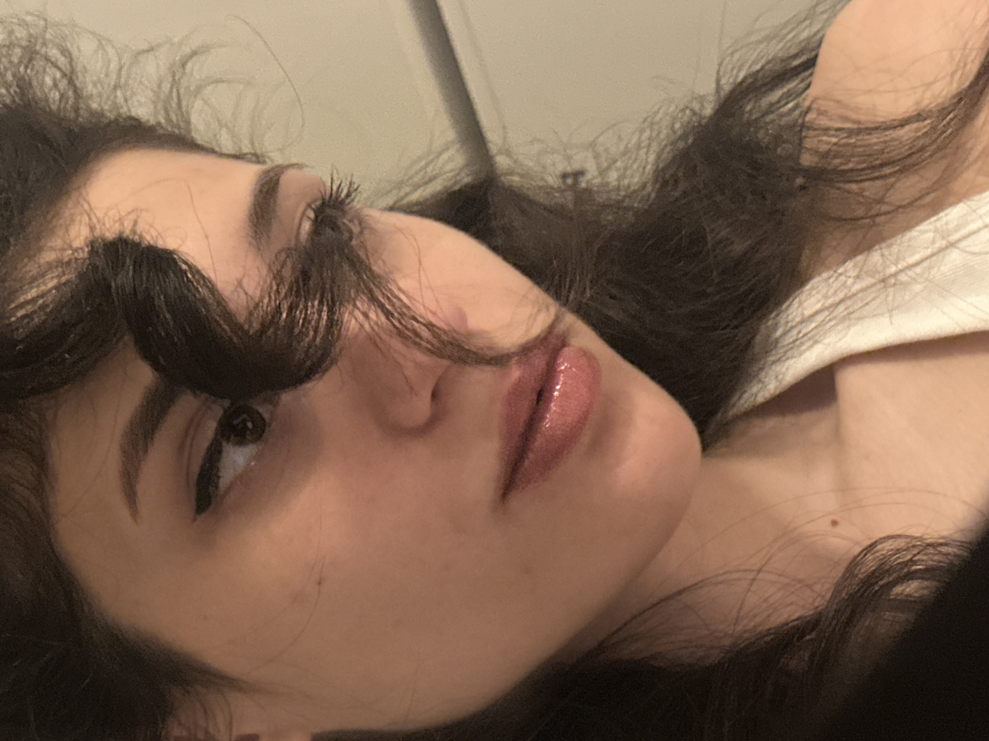 A close-up, slightly angled portrait shows the left side of a young woman's face and upper torso. Her dark hair is styled in loose waves, with a section framing her eye and falling across her cheek. She is wearing a white top, the strap of which is visible. The woman's skin appears smooth, and she has light makeup, including eyeliner and lip gloss. Her eyes are looking downwards and to her left. The background is a plain, light-colored wall, with a horizontal grey bar visible in the upper right corner. The overall lighting is soft and warm. There are no identifiable landmarks or objects within the frame that would suggest a specific location in Sofia, Bulgaria.