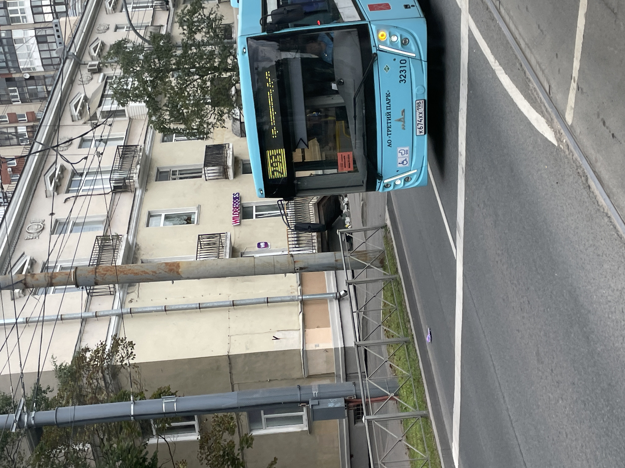 A light blue bus with the number 32310 is traveling on an asphalt road in front of a building. The bus has a digital display on its front showing the number 255 and some Russian text. The building behind the bus is a multi-story structure with cream-colored walls and balconies. There are trees and utility poles along the street. A silver metal fence runs along the side of the road. There is a white pedestrian crossing in the foreground. The license plate on the bus reads "K674XX 198".