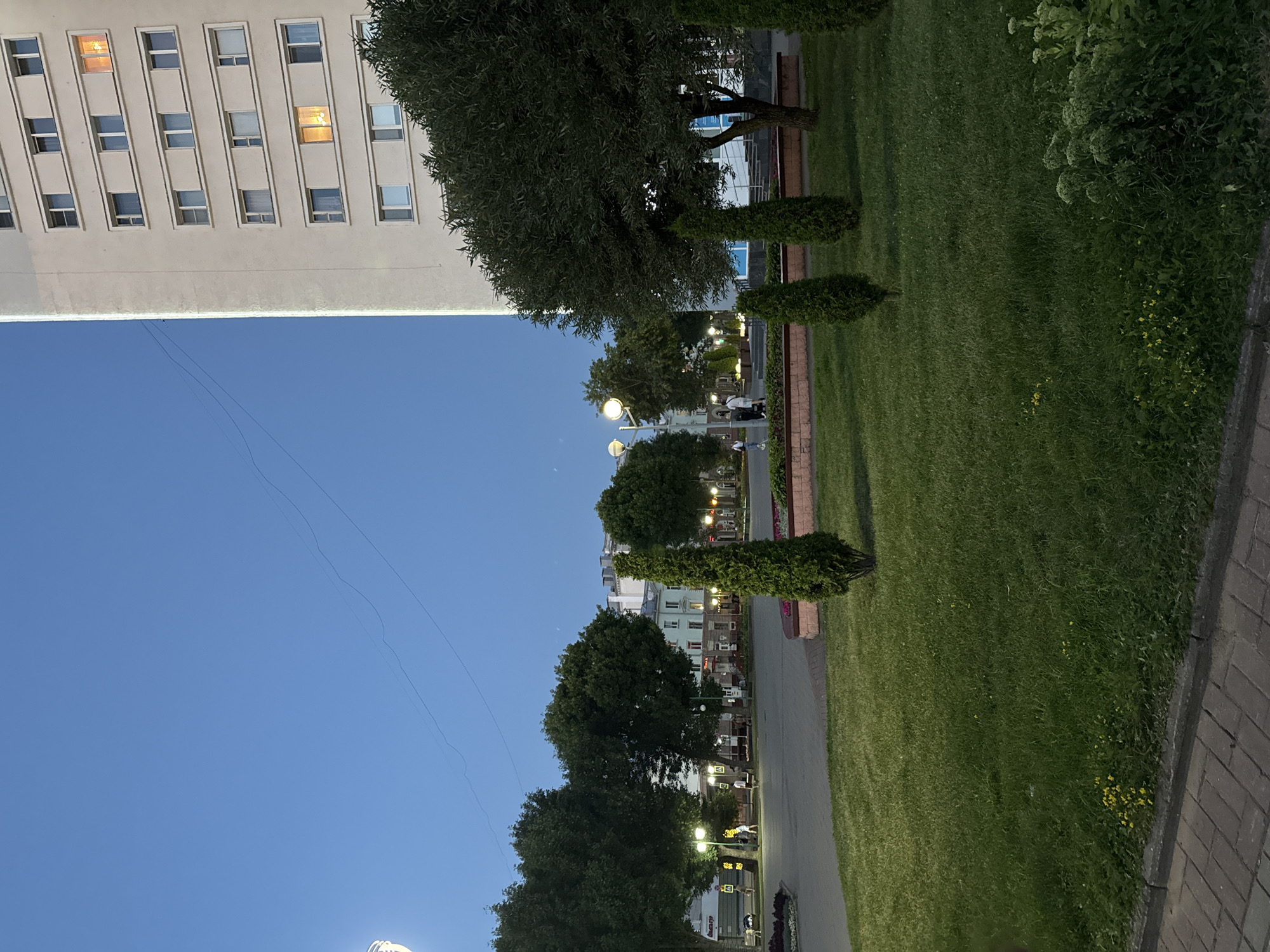 The image depicts an urban scene in Mahilyow, Belarus, at dusk or early evening. In the foreground, a well-maintained lawn with small yellow wildflowers borders a brick walkway. Several conical evergreen trees are planted in the lawn. In the middle ground, a paved area features lampposts with glowing orbs, illuminating trees and the facade of buildings across the street. A few individuals are walking in this area. The background is dominated by a tall, light-colored apartment building on the right, its windows mostly dark, though a few show illuminated interiors. To the left of the apartment building, more trees and buildings are visible. The sky above is a deep blue, indicating the transition from day to night, with faint power lines stretching across it. The overall impression is of a quiet city square as evening sets in.