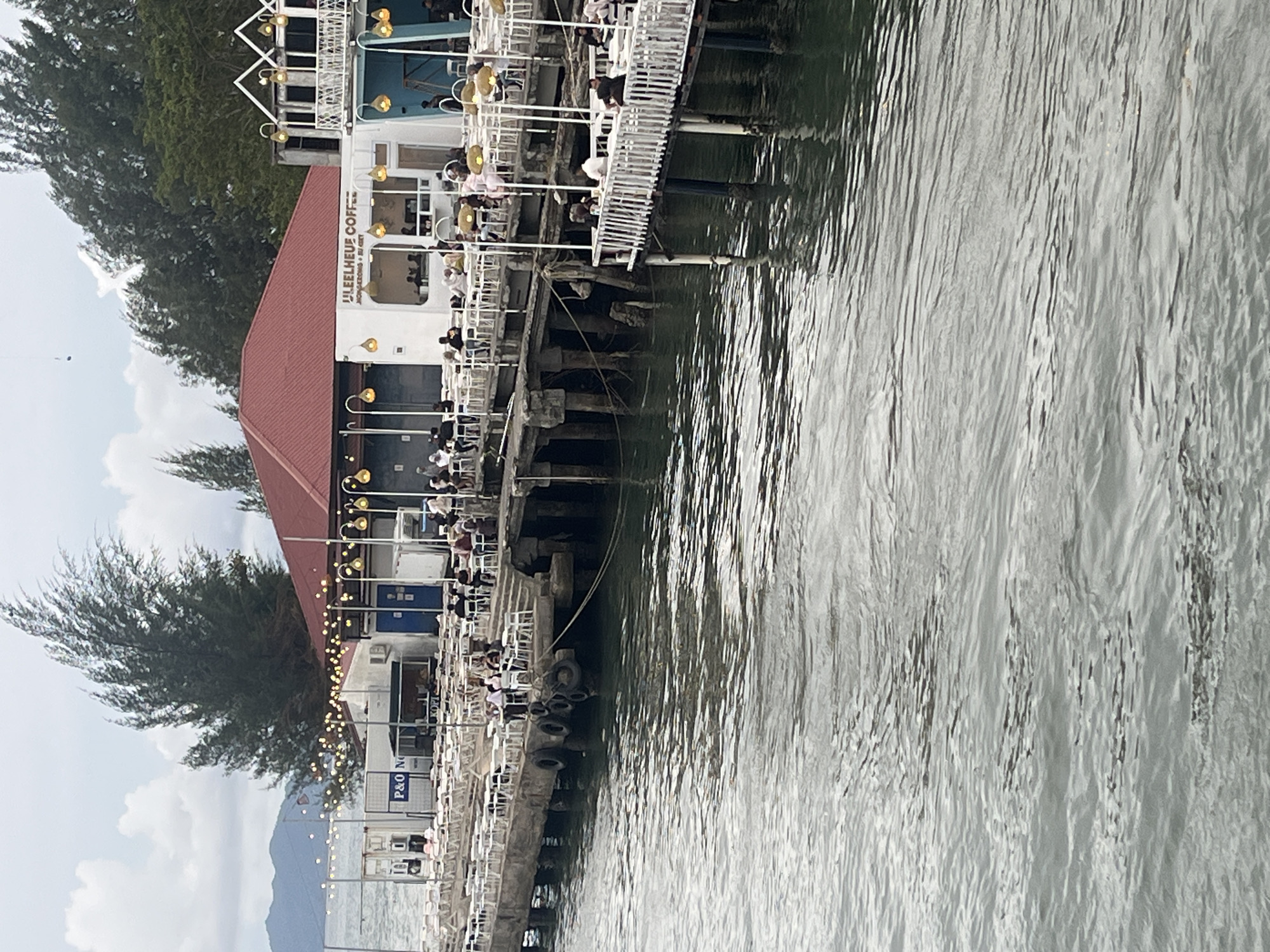 The image depicts a waterfront cafe and seating area on a pier in Banda Aceh, Indonesia. Several people are seated at tables on the pier, some of whom appear to be patrons of the cafe. The cafe itself is a white building with blue accents and features signage that reads "ULEELHEUS COFFEE." String lights are visible overhead, illuminating the seating area. The pier extends into the water, which appears choppy. In the background, trees and mountains can be seen. The overall scene suggests a bustling outdoor dining environment by the sea.