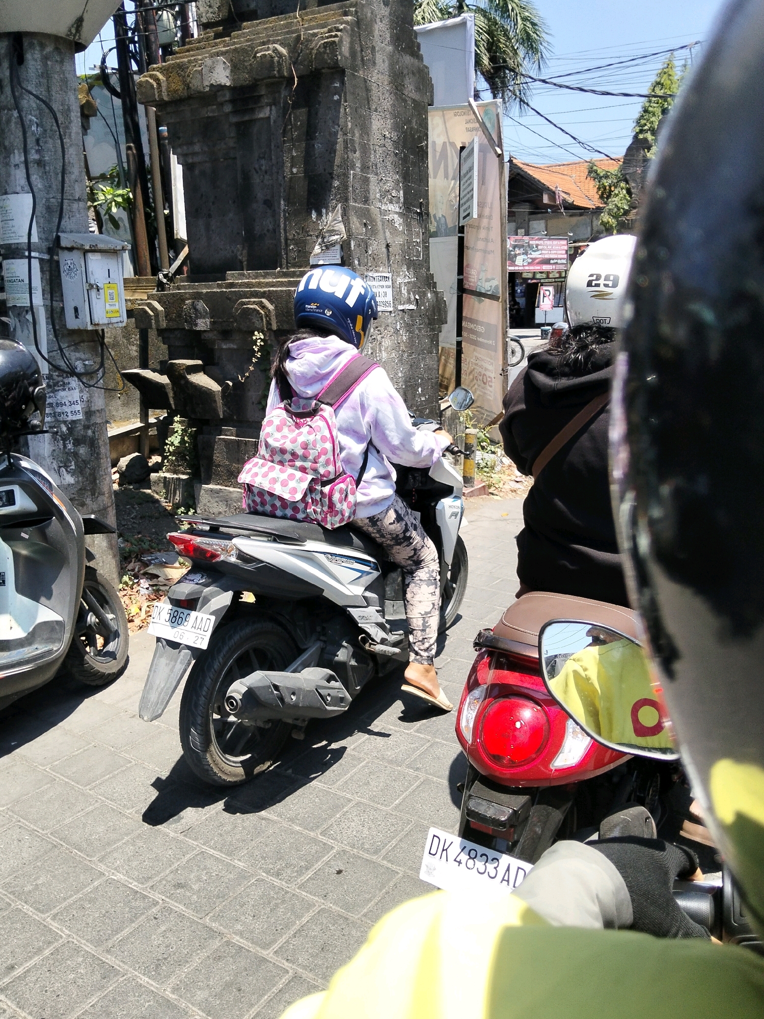 A person wearing a blue helmet and a light pink hoodie is riding a white scooter on a cobblestone street in Denpasar, Indonesia. The person is carrying a pink backpack with white cherry patterns. Another person, partially visible, is wearing a black hoodie and riding a red scooter in front of the white scooter. The red scooter has a license plate that reads "DK 4833 AD". The environment appears to be a city street with buildings, trees, and power lines. The lighting suggests it is daytime.