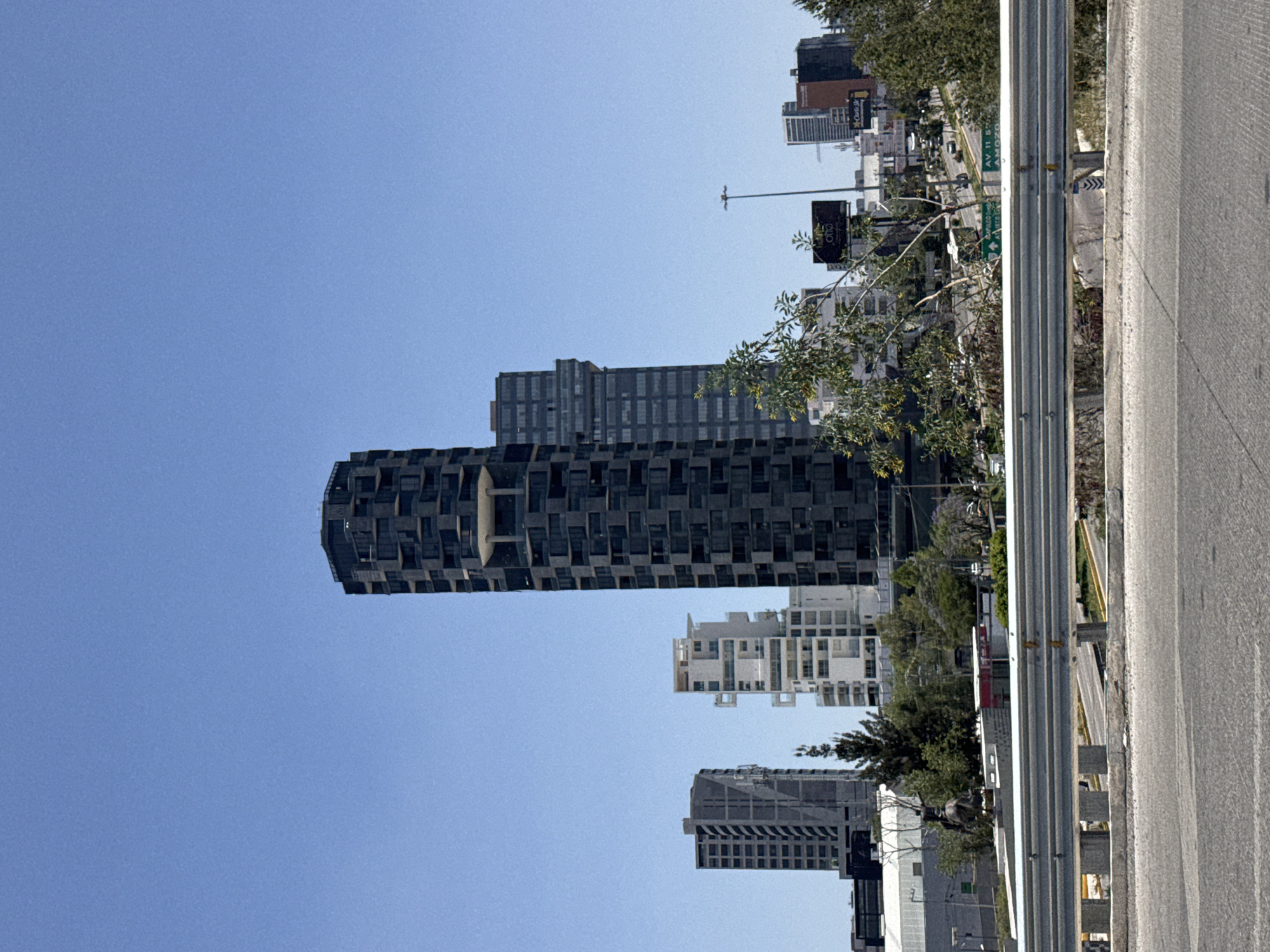 The image shows a view of several buildings in San Bernardino Tlaxcalancingo, Mexico, from a roadside perspective. A tall, dark building with a rounded shape and a repeating pattern of rectangular openings dominates the middle ground. To the right are a few taller, more conventional high-rise buildings. A highway runs along the right side of the frame, with a metal guardrail separating it from the foreground. The sky is a clear, bright blue. There are trees and signs visible among the buildings.