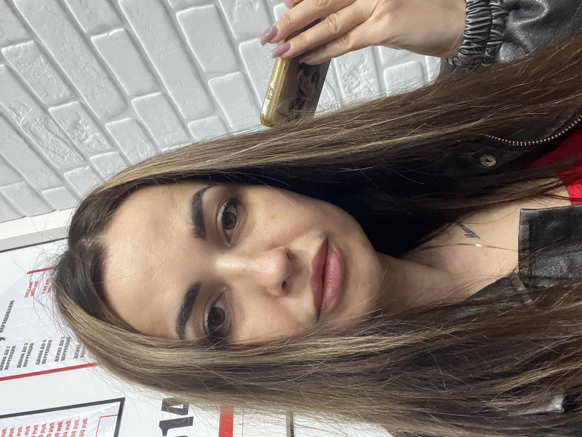 A female adult with long, straight, brown hair with lighter highlights, brown eyes, and a neutral expression is captured in a head-and-shoulders selfie. She is wearing a black leather jacket, under which a red garment is partially visible, along with a thin gold chain necklace and a dark text tattoo on her upper right chest. Her right hand, featuring manicured nails with light purple polish, holds a gold-colored smartphone near her right ear, with the screen facing the camera.
The background consists of a white wall. To the left, two vertical signs display text in Russian, including words like "НАРАЩИВАНИЕ" (nail extension) and "КОРРЕКЦИЯ" (correction), alongside prices in rubles (e.g., 2000, 2700). To the right, a white textured wall mimics brickwork. The scene is an indoor commercial setting, likely a beauty salon or similar establishment, in the city of Unknown, Unknown.