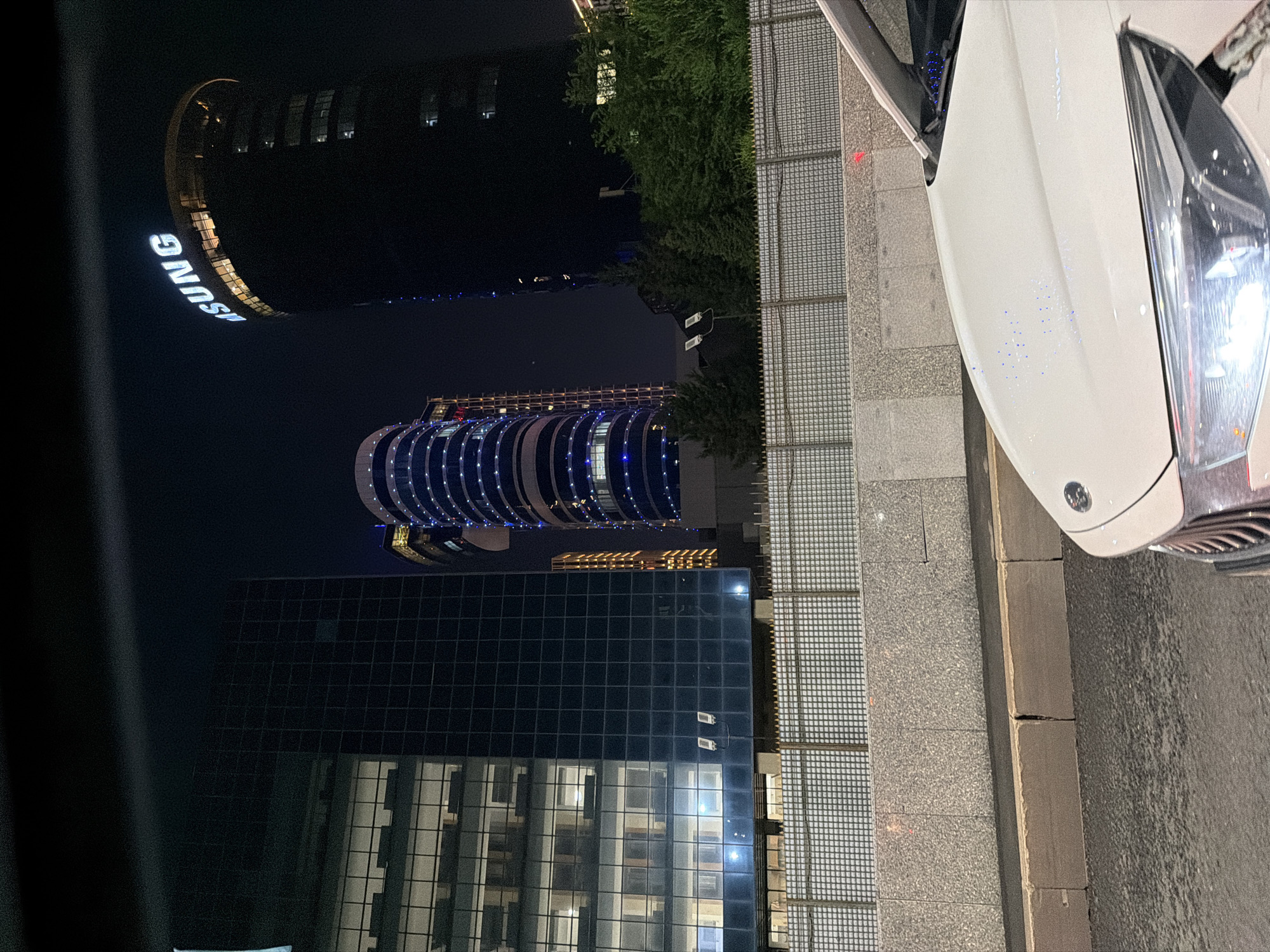 This is a night-time exterior shot of modern skyscrapers in Astana, Kazakhstan. The foreground features the hood of a white car, with its headlights illuminated. A grated fence runs horizontally across the middle ground, separating the road from a concrete barrier and then the buildings. The buildings are tall and appear to be made of glass and metal. One prominent building on the right displays a large illuminated "SAMSUNG" logo. Other buildings have patterned lighting, with some featuring vertical stripes of blue lights. Green trees are visible behind the fence, in front of some of the buildings. The sky is dark, indicating it is nighttime. The scene suggests a bustling urban environment with contemporary architecture.