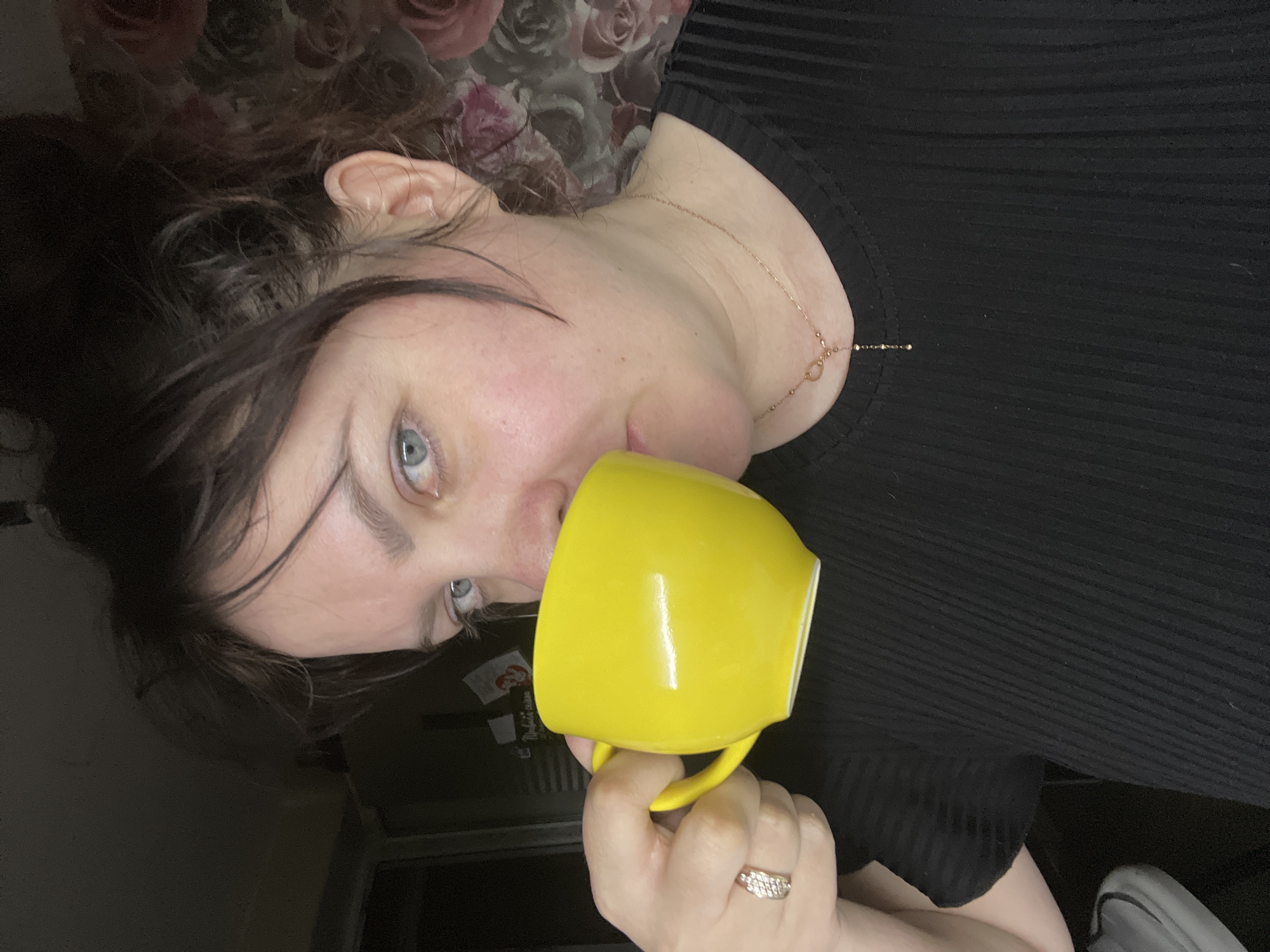 An adult female is depicted in a close-up, chest-up shot, drinking from a bright yellow ceramic mug. She possesses fair skin, blue eyes, and dark brown hair, which appears to be styled loosely or tied back. She is wearing a black, short-sleeved top with a ribbed texture and a thin gold-colored necklace featuring a circular charm and a dangling element. A ring with small, clear stones is visible on her right ring finger as she holds the mug to her mouth. The background is dimly lit, with a dark, indistinct area to her left and a section of dark floral wallpaper with large, muted pink roses to her right. The scene is located within an interior setting in the city of Noviy, Russia.