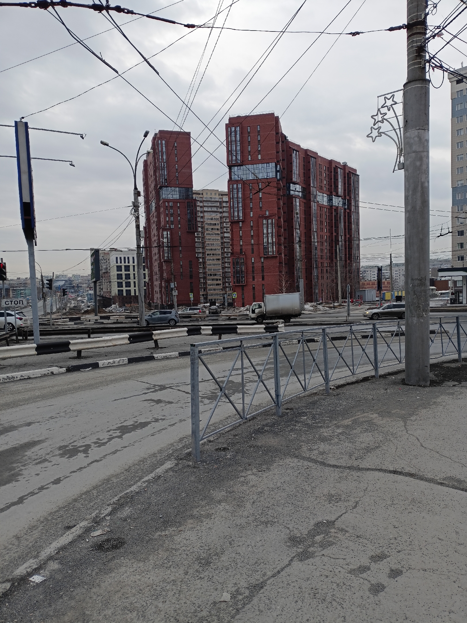 The image depicts a street scene in Novosibirsk, Russia, dominated by two tall, red-brick buildings with a modern design. The buildings appear to be residential, with visible rows of windows. A multi-lane road separates the viewer from these buildings, with a gray metal fence running along the side of the road. Cars and a truck can be seen driving on the road. Overhead, there is a network of power lines and cables crisscrossing the sky. The sky is overcast, suggesting a cloudy day. In the foreground is a gray, somewhat worn sidewalk or shoulder of the road. A streetlight pole stands to the right, adorned with a decorative star-shaped light fixture. Other buildings are visible in the background. A road sign with the word "СТОП" (STOP) is visible on the left.