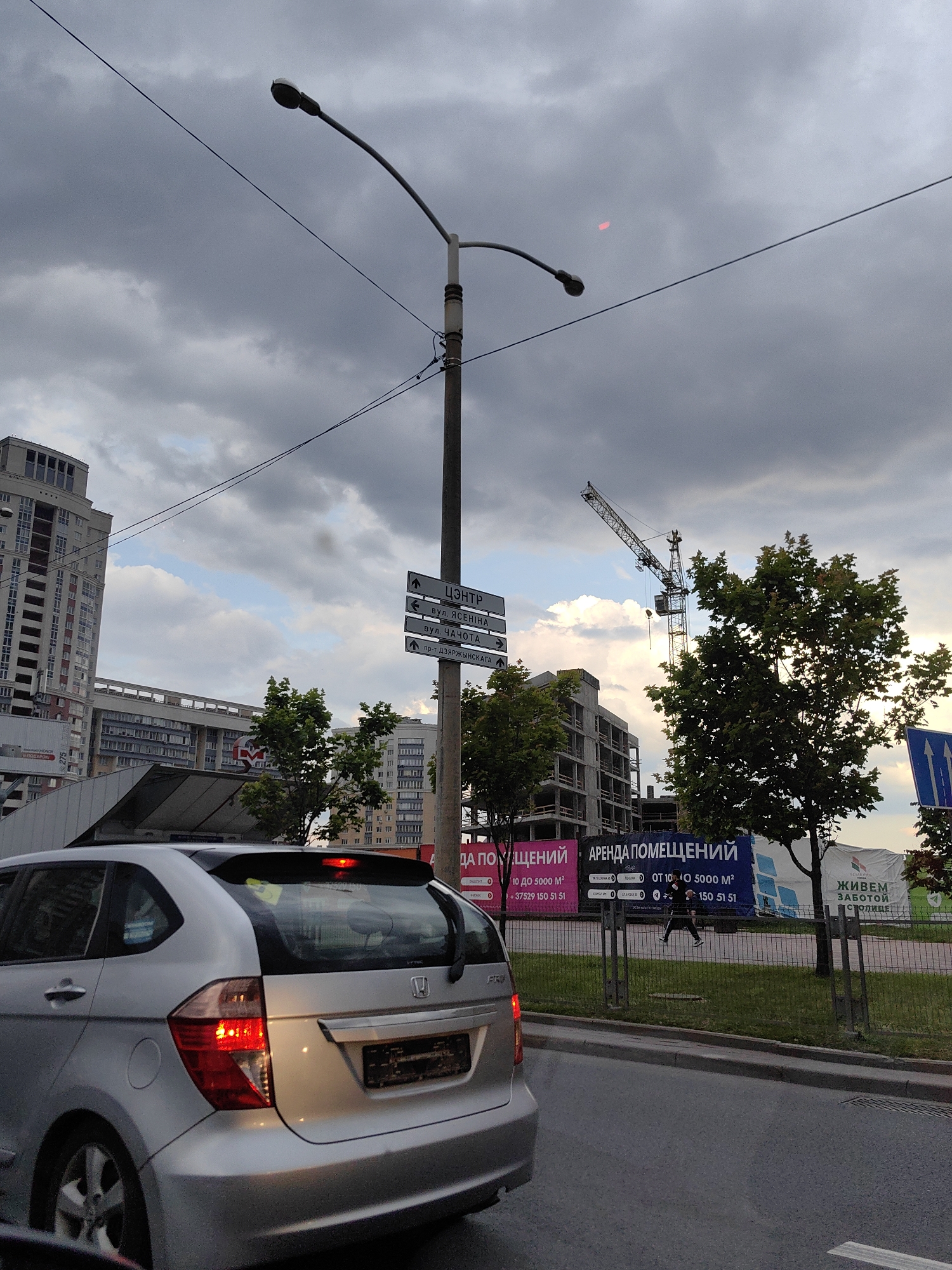 The image shows a street scene in Minsk, Belarus, during the daytime under a cloudy sky. In the foreground, the rear of a silver Honda Fit is visible, with its brake lights illuminated. The car is stopped at what appears to be an intersection or traffic light.
Behind the car, a tall street lamp with a curved arm and a single light fixture stands prominently. Attached to the pole of the street lamp are directional signs written in Cyrillic script. One sign indicates "ЦЕНТР" (Center), with an upward arrow. Below it are signs pointing to "вул. ЯСЕНІНА" (Yasenin Street) and "вул. ЧАХОТА" (Chakhota Street), each with a rightward arrow. A further sign points to "пр-т. ДЗЯРЖЫНСКАГА" (Dzerzhinsky Avenue) with an upward arrow.
In the middle ground, several modern buildings are visible. To the left, a multi-story residential building with a facade of white and red elements dominates the skyline. Further to the right, a construction site with a crane is active, with concrete structures of a building under construction. Large billboards are displayed in front of the construction site. One billboard reads "АРЕНДА ПОМЕЩЕНИЙ" (Rent Premises) with additional text and contact information. Another billboard shows the text "ЖИВЕМ ЗАБОТОЙ СТОЛИЦЕ" (We live with the care of the capital).
On the right side of the image, a pedestrian, dressed in dark clothing and running shoes, is seen on a sidewalk behind a metal fence, moving from left to right. The street is paved with asphalt, and a grassy area with trees is present along the sidewalk. The overall atmosphere suggests an urban environment with ongoing development.