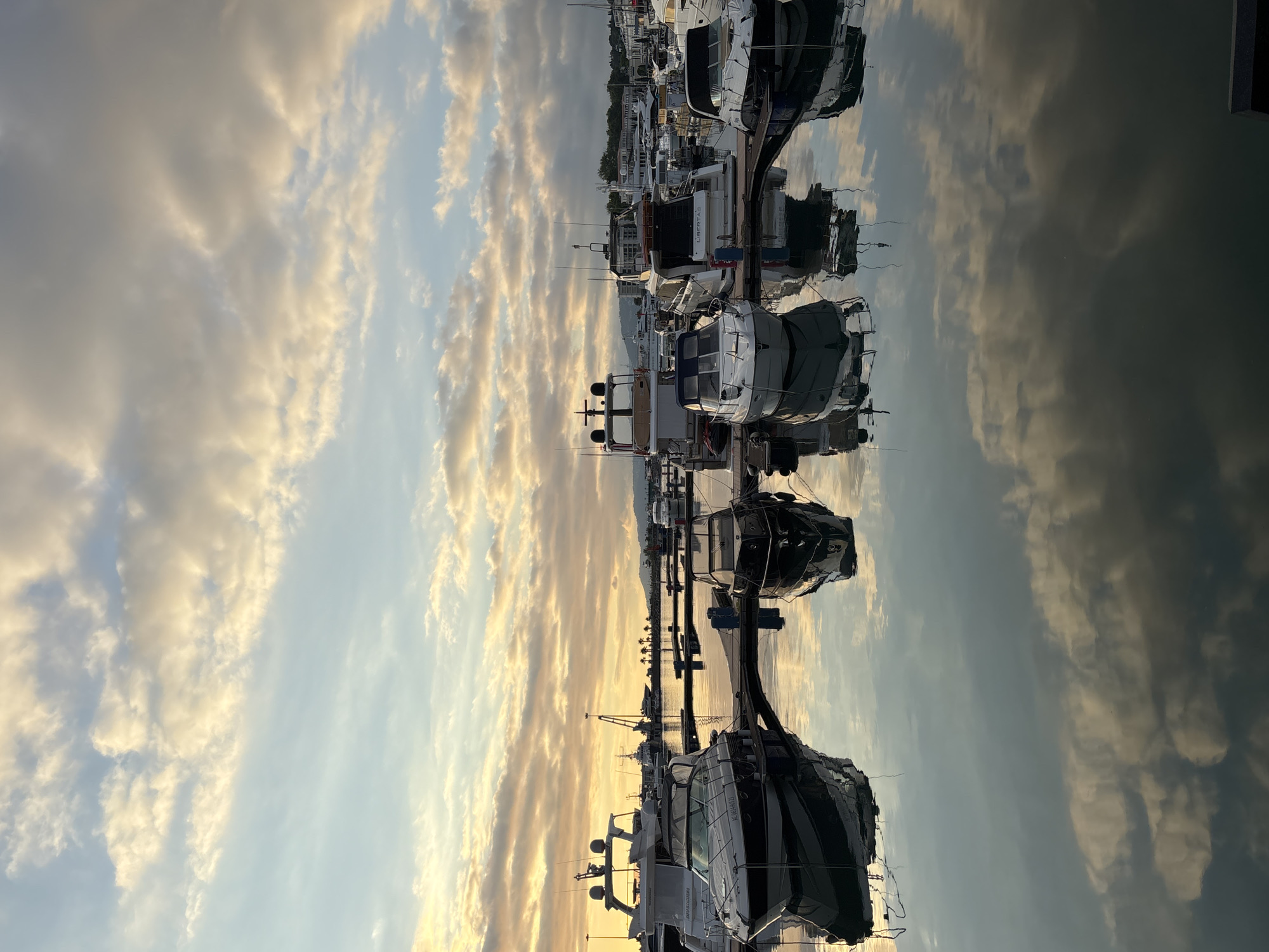 The image depicts a marina filled with various yachts and boats docked along a pier. The scene is illuminated by the soft, warm light of either sunrise or sunset, casting a golden glow on the scattered clouds. The sky is a blend of blue and pale orange, with streaks of white and yellow clouds. The water is calm, reflecting the sky and the boats, creating a mirror-like effect.
Several yachts are prominently visible, with their sleek designs and dark hulls contrasting against the lighter surroundings. Some boats are covered with tarpaulins, suggesting they are either being stored or are temporarily out of use. The pier structure is visible, with mooring lines and other nautical equipment. In the background, a line of palm trees and buildings can be seen along the coastline, hinting at a temperate climate. The overall atmosphere is serene and peaceful, with the natural beauty of the sky and water complementing the man-made structures of the marina.
Visible text includes "LIBERTAS" on the side of one of the yachts and "AX6949AU" on another. The location context is Sochi, Russia.