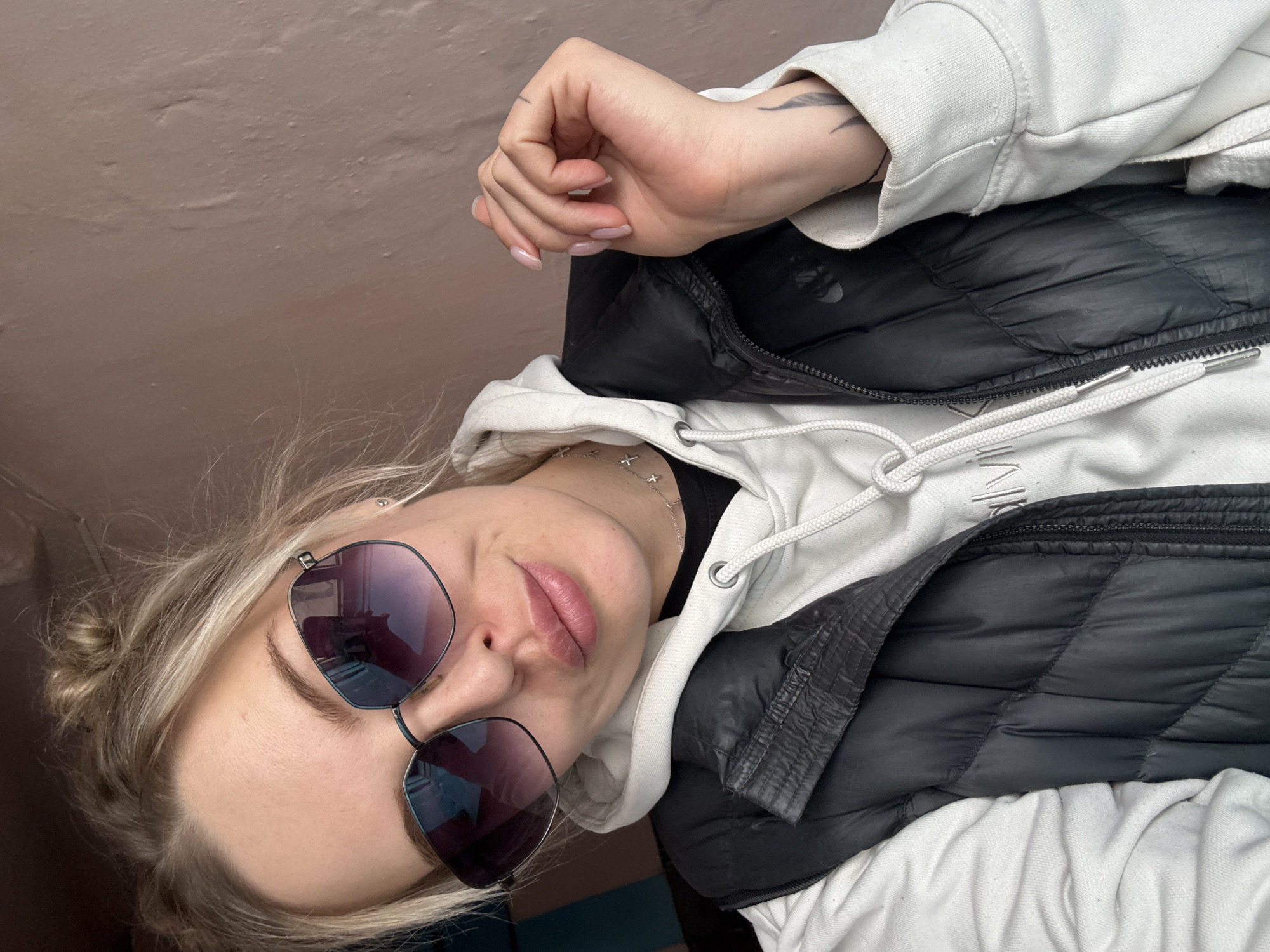 An indoor, close-up portrait of a young woman, rotated 90 degrees counter-clockwise. She has light blonde or light brown hair, styled in a loose bun on one side, and wears large, reflective dark sunglasses. She is dressed in a light-colored hooded sweatshirt, with the text "CALVIN KLEIN" faintly visible on the front, layered with a black puffer vest over a dark top. A silver necklace adorned with small cross charms is visible around her neck. Her lips are slightly parted, and she has a neutral, relaxed expression. Notable details include light pink nail polish on her fingernails and a subtle feather-like tattoo on her inner forearm. The background is a plain, light brown textured wall. The photo captures a moment of casual posing, likely a selfie, within what appears to be an interior space in Borzya, Russia, under ambient lighting.