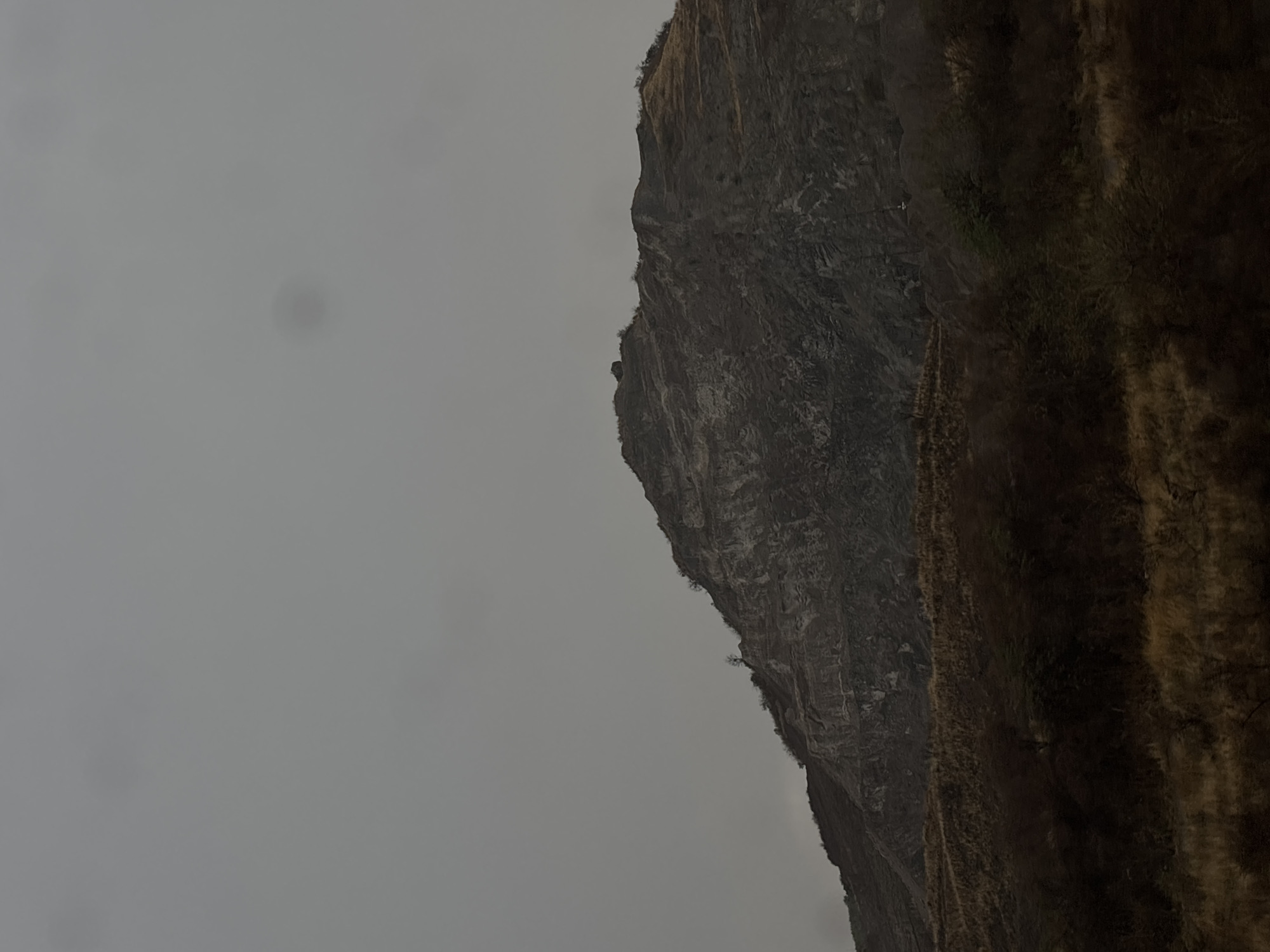 The image depicts a mountainside, presumably near San Bernardino Chalchihuapan, Mexico, under an overcast sky. The left portion of the frame is filled with a gray, featureless sky, while the right side showcases the steep, rugged terrain of the mountain. The mountain's surface is varied, exhibiting areas of dark, rocky cliffs alongside patches of brown vegetation. The overall environment appears harsh and potentially remote. There are no people, animals, or man-made structures visible in the image.