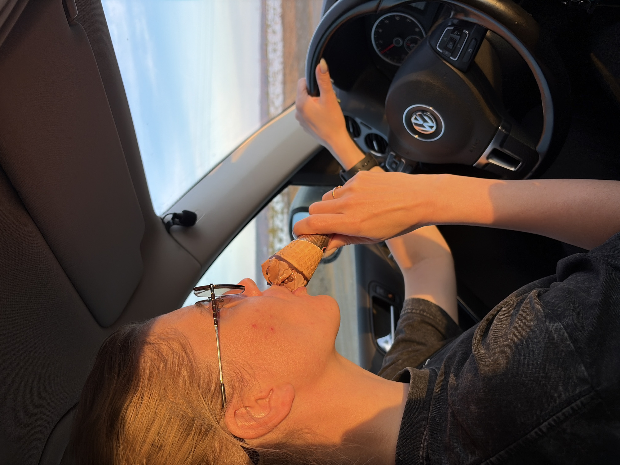 A woman wearing glasses is in the driver's seat of a Volkswagen, eating an ice cream cone. The sky is visible through the windshield, suggesting it is daytime. The location is unknown and cannot be determined from the image.