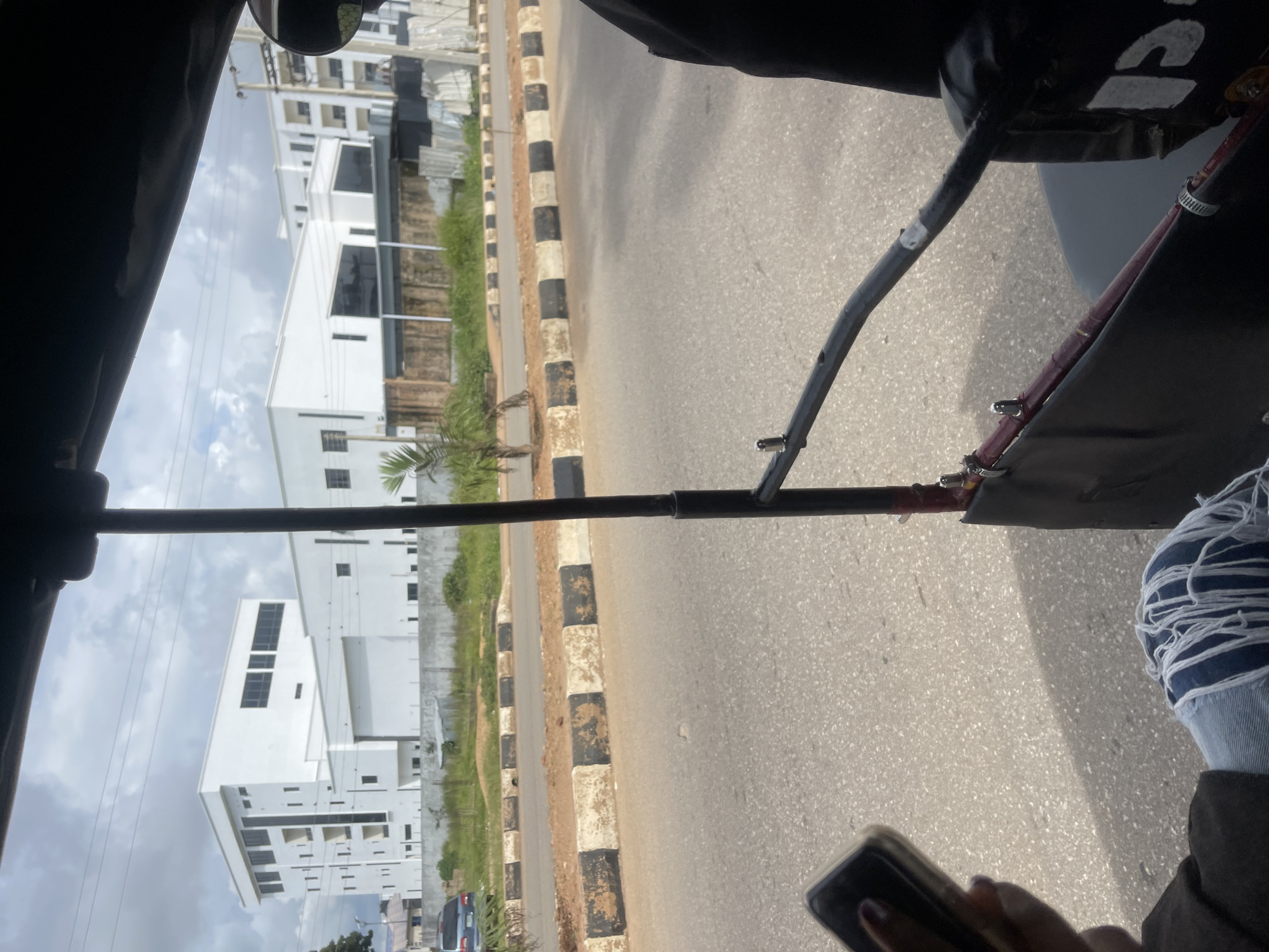 The image shows a view from inside a vehicle, likely a tuk-tuk, looking out at an urban environment in Bar Jirgi Summa, Nigeria. Modern white buildings with numerous windows dominate the background. A paved road with a black and white striped curb separates the road from a grassy area in front of the buildings. A few small trees and some construction materials are visible in the grassy area. The foreground shows a person's hand holding a smartphone and their legs wearing ripped jeans. The overall scene depicts a typical street view with contemporary architecture under a cloudy sky.