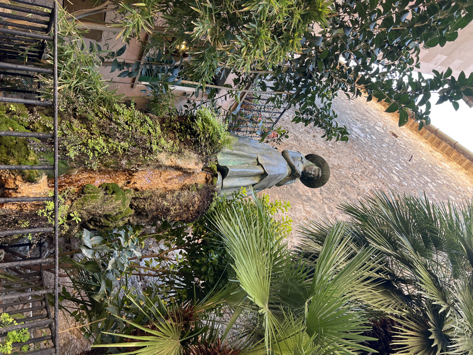 A bronze statue of a female figure, positioned centrally, dominates the scene in Valldemossa, Spain. The statue depicts a woman wearing a wide-brimmed hat and a long dress or apron, with her head inclined downwards and her hands held together at chest level, suggesting contemplation or work. She stands atop a naturalistic fountain structure composed of dark, textured rock heavily encrusted with green moss and small plants, with visible streams of water trickling down its surface.
The environment is characterized by dense, vibrant green foliage surrounding the statue. Large, broad-leafed trees and bushes frame the left side, while prominent palm fronds are visible on the right. In the background, a tall, weathered stone wall with a visible mortar pattern rises, culminating in a terracotta tiled roofline at the top. To the immediate left of the statue, a segment of a multi-story building with a light peach-colored facade is visible, featuring a dark window and a black wrought-iron balcony railing. A black metal fence or railing runs horizontally across the lower foreground, partially obscuring the base of the fountain and the ground level. The scene is illuminated by natural light, casting distinct shadows and highlighting the various textures of the stone, metal, and organic elements. No living persons are present.