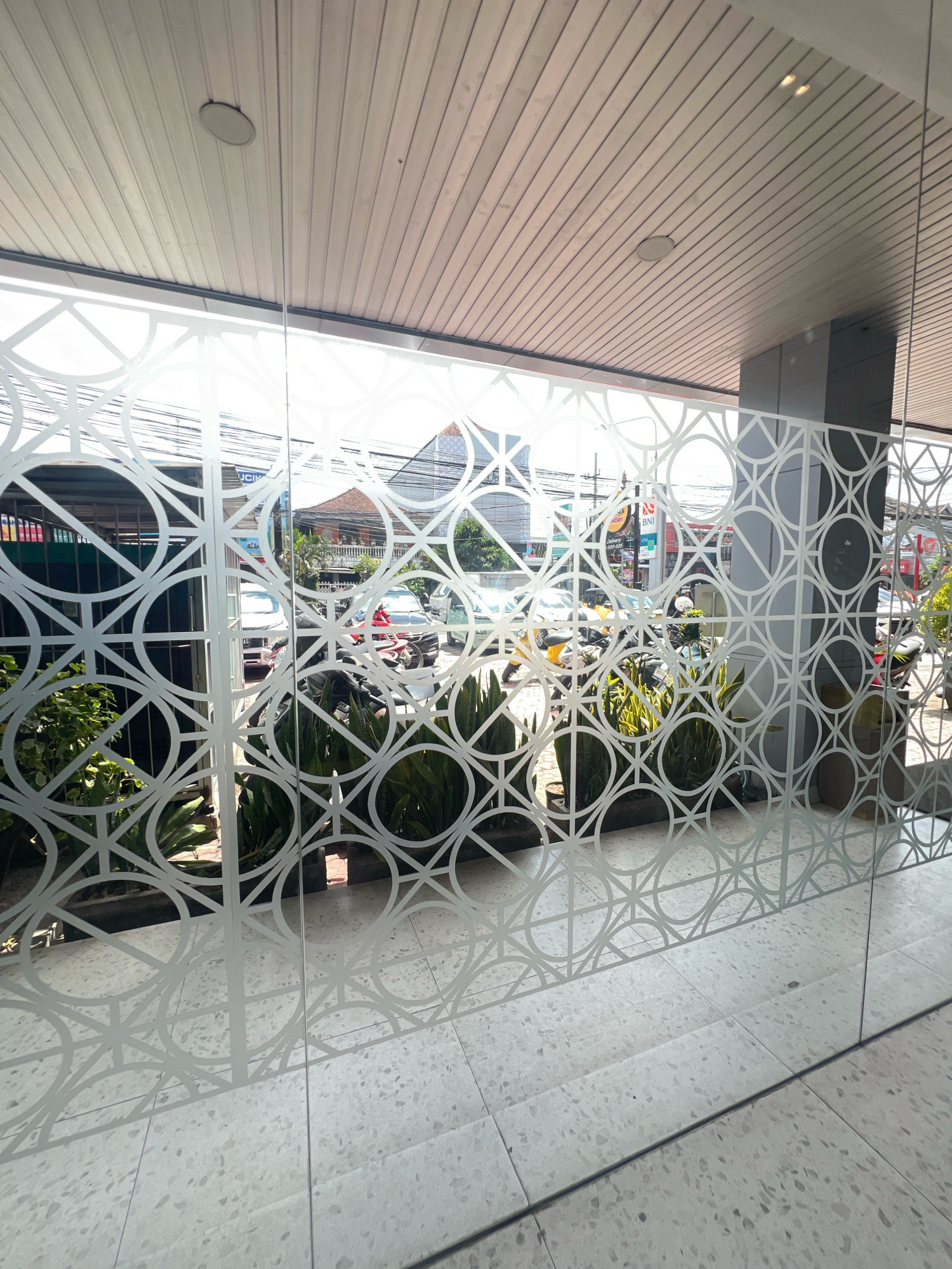 The image shows a street view in Denpasar, Indonesia, as seen through a glass wall with a decorative geometric pattern. The pattern consists of interlocking circles and squares. Outside the glass, there are parked vehicles, including cars and motorcycles, and some potted plants. The ceiling appears to be made of wooden panels. The floor is tiled with a speckled terrazzo pattern. The overall scene suggests an urban environment on a bright, sunny day.