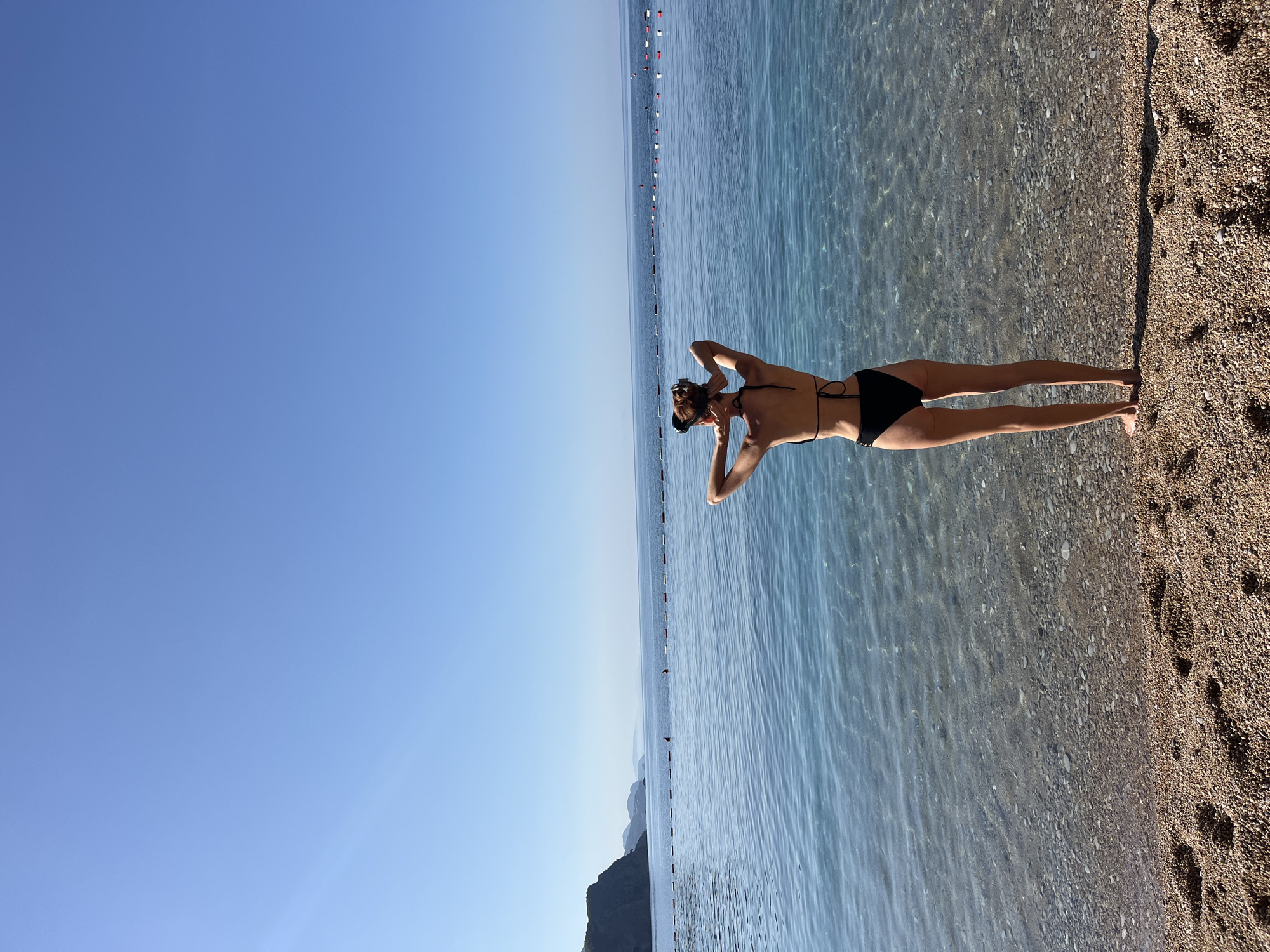 A high-angle, full-shot photograph depicts a female subject standing on a pebbled beach, facing away from the viewer and looking out at the sea under a clear blue sky.
The subject is a slender adult female, positioned centrally, wearing a black two-piece bikini. Her arms are raised, with her hands clasped behind her head. A black snorkeling mask rests on top of her head. Her back, arms, and legs are visible.
The immediate foreground is a coarse pebbled beach, composed of various shades of brown and grey stones. Indentations consistent with footprints are visible in the pebbles directly behind the subject. A distinct, elongated shadow extends from the subject's feet towards the right side of the frame, indicating direct sunlight from the left.
The midground features calm, clear water. The pebbled seabed is visible through the shallow, transparent water near the shore. Ripples are present on the water's surface, reflecting the bright sky.
Further out, the water transitions to a deeper blue. A linear arrangement of buoys, alternately colored red and white and connected by a rope, spans across the visible expanse of the sea, demarcating a swimming area. Two additional, more distant red pyramidal buoys are discernible beyond this line.
The background consists of a clear horizon line separating the sea from an uninterrupted, vivid blue sky, devoid of clouds. To the far left, a rugged, mountainous coastline with prominent cliffs and receding hills is visible, extending into the distance and appearing somewhat hazy.
This scenery is consistent with coastal areas of Budva, Montenegro, characterized by pebbled beaches, clear Adriatic waters, and a mountainous hinterland.