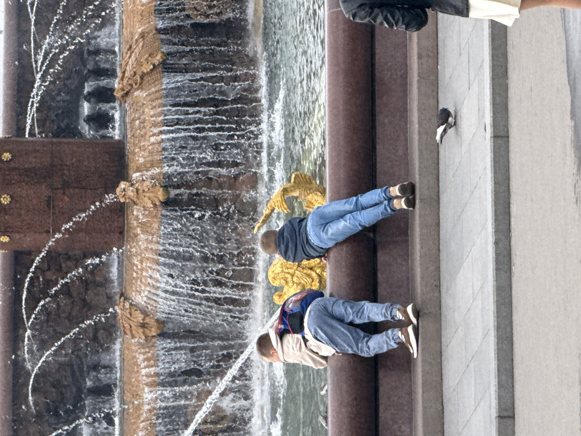 The image shows a close-up view of a fountain in a public area, likely Moscow, Russia. Two young boys are standing on the edge of the fountain, looking out at the water. One boy is wearing a light-colored jacket and blue jeans, with a backpack on his back. The other boy is wearing a dark hooded jacket and blue jeans. A pigeon is seen on the pavement in front of the fountain. In the background, the fountain is depicted with water cascading down, and decorative elements, possibly made of gold, are visible. The overall scene suggests a recreational or tourist location.