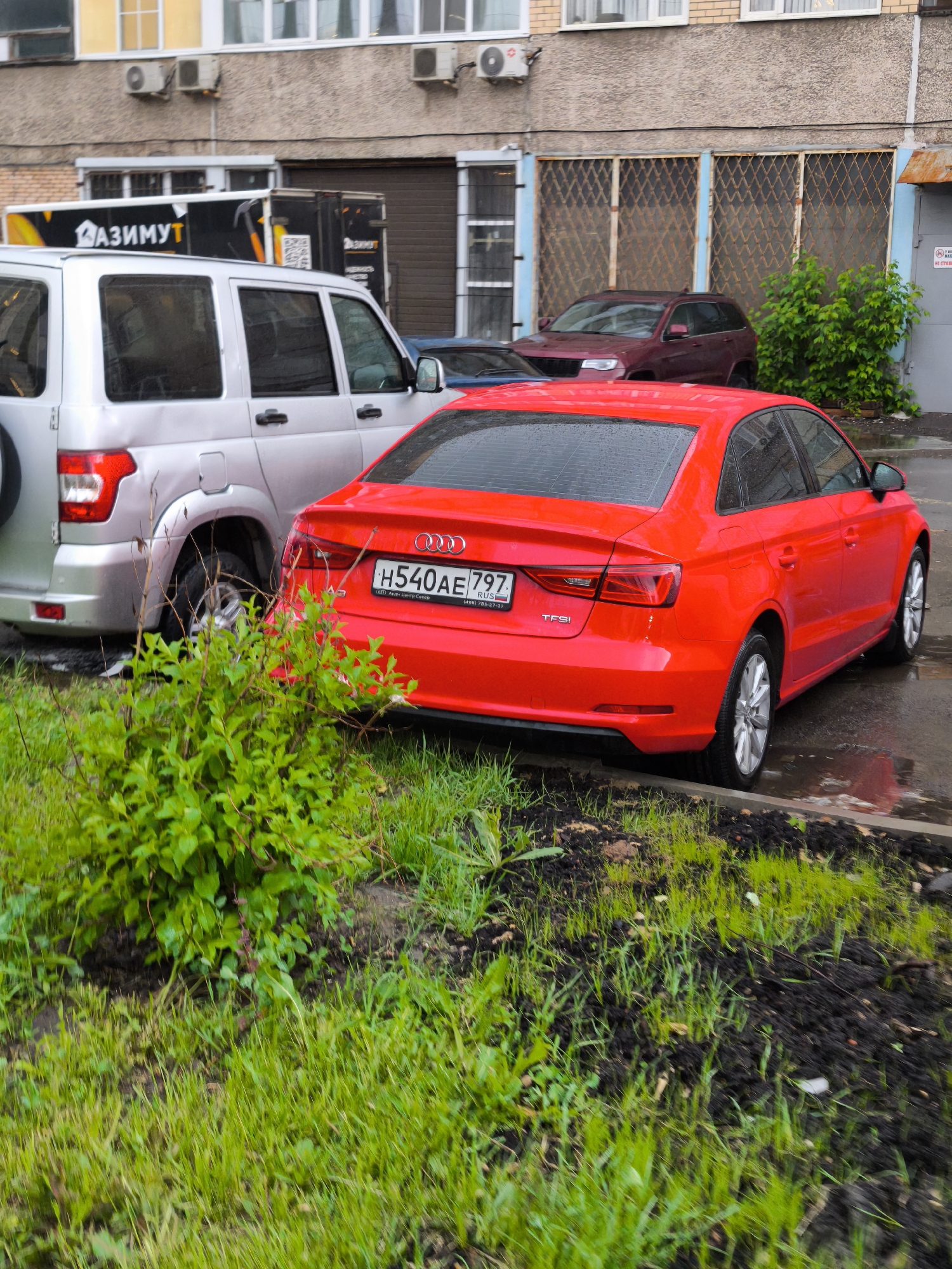 The image shows a red Audi A3 parked on the side of a street in what appears to be an urban area, possibly Moscow, Russia, as indicated by the Russian license plate. The car is parked next to a silver SUV, and behind it is a maroon SUV. The ground is a mix of grass and dark soil, suggesting a small patch of vegetation next to the road. Behind the cars is a building with visible air conditioning units on the facade. There is a box truck with the word "КАЗИМУТ" visible. The overall setting seems to be a residential or commercial area.
