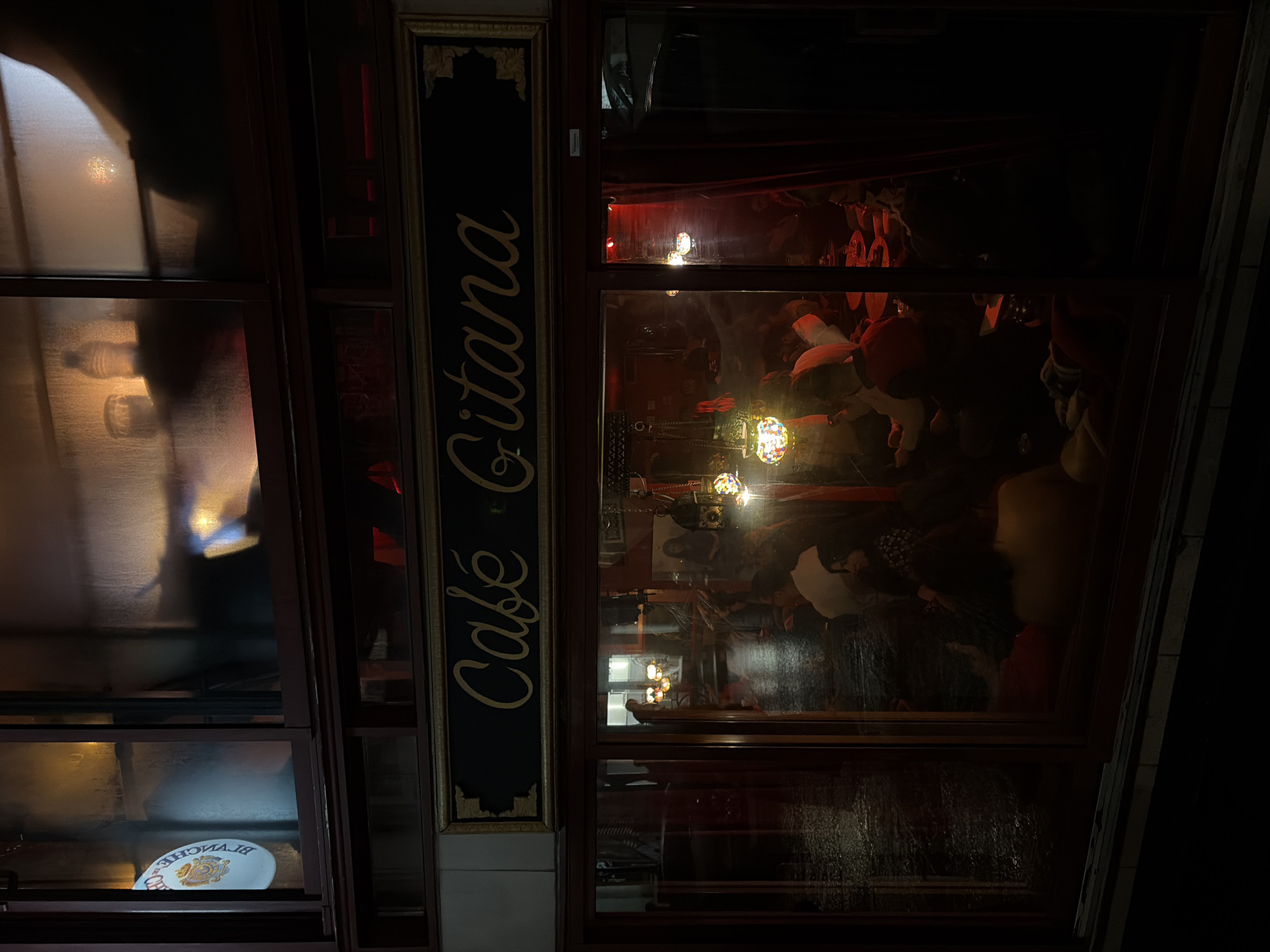 A nighttime view captures the facade of "Café Gitana" in Montreal, Canada. The café's name is written vertically on a narrow, dark sign bordered with a gold-toned trim, positioned between window displays. Through the windows, the interior is visible with blurred figures suggesting patrons inside, lit by warm-toned light fixtures. The reflections on the glass indicate a wet surface. The immediate surroundings are dark, emphasizing the light emanating from within the café. A shelf in the window displays a plate with branding on it.