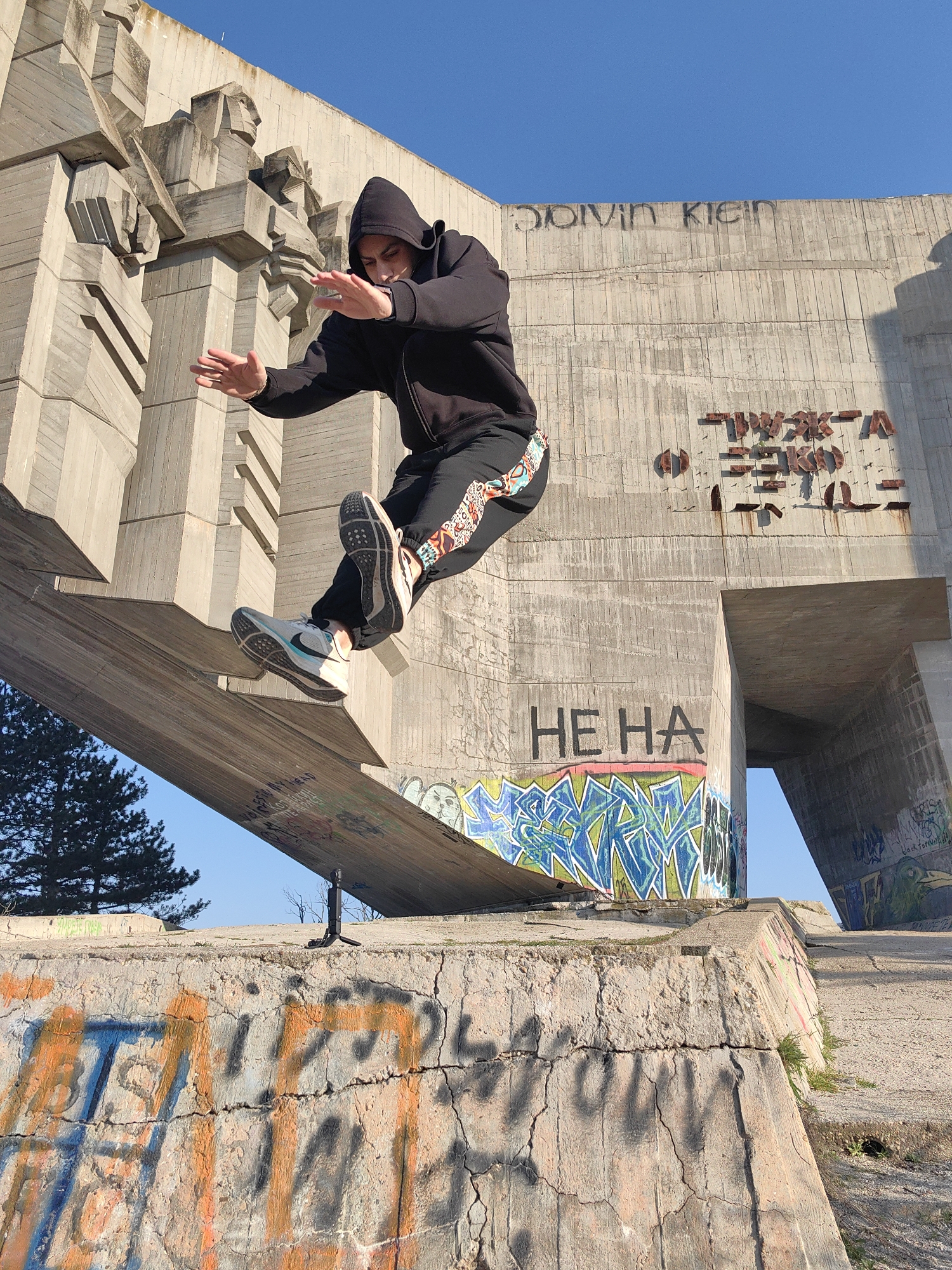 The image shows a person in a black hooded sweatshirt and patterned sweatpants jumping from a concrete structure. The person wears grey and white running shoes. The background is a large, brutalist-style concrete monument with graffiti. "HE HA" is written in black letters. "Calvin Klein" is also written at the top of the structure. The location is the Monument of the Bulgarian-Soviet Friendship in Varna, Bulgaria. The sky is clear blue. A small tripod stand is visible.