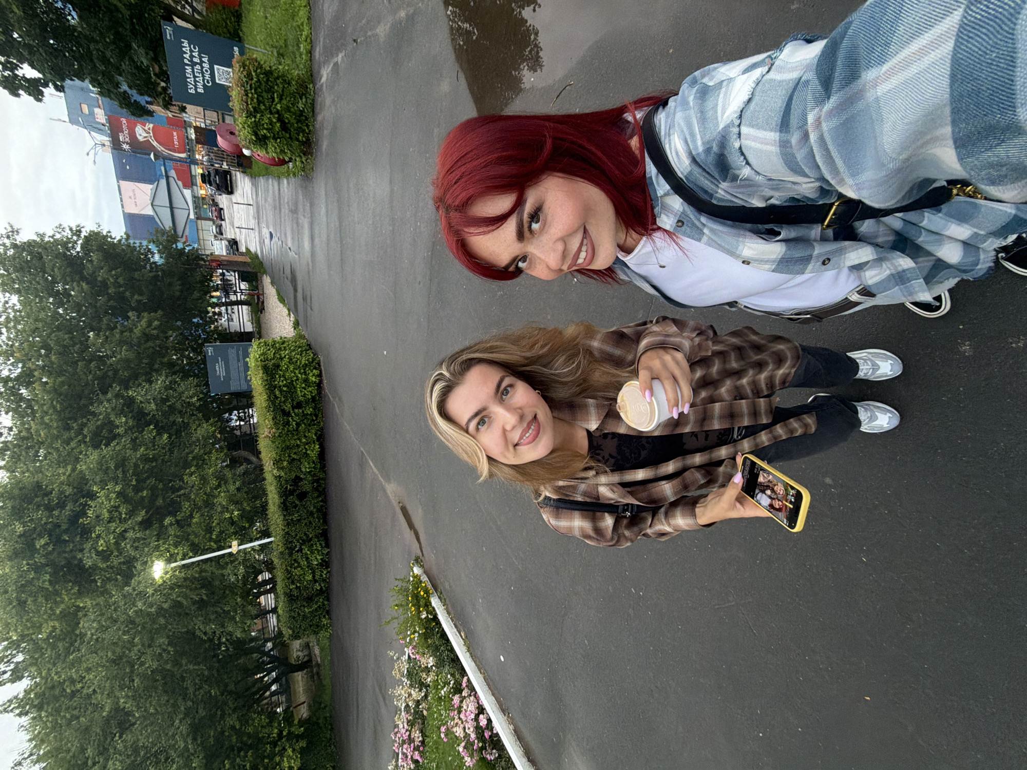 Two young women are posing for a selfie outdoors, with a blurred background of trees and buildings. The woman in the foreground has blonde hair and is wearing a brown and black plaid shirt over a black top. She is holding a cup in her right hand and looking at the camera with a smile. She is also holding a smartphone in her left hand, displaying a group photo. The woman behind her has long, red hair and is wearing a blue and white plaid shirt over a white top. She is also smiling at the camera. The setting appears to be a paved walkway or road, with some greenery and a lamppost visible. The overall mood is casual and friendly. Visible text includes "БУДЕМ РАДЫ ВИДЕТЬ ВАС СНОВА!" on a sign in the background, which translates to "WE WILL BE GLAD TO SEE YOU AGAIN!"