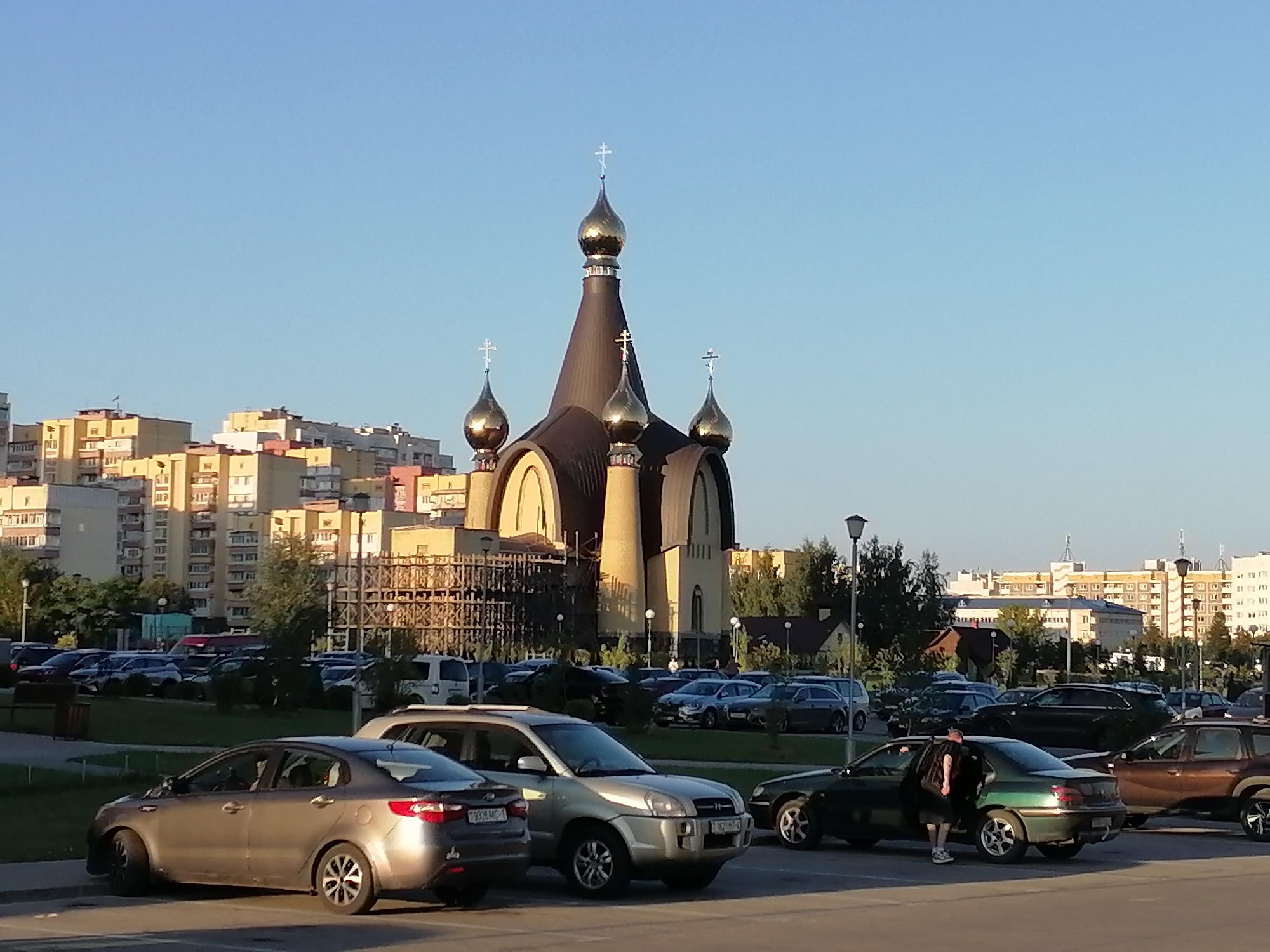 This image shows an outdoor scene in the city of Hrodna, Belarus. In the foreground, a silver Kia Rio is parked facing left. To the right of the Kia Rio, a silver Hyundai Tucson is parked facing the viewer. A person wearing a black t-shirt and shorts is exiting a dark green Peugeot 407 sedan, which is parked to the right of the Hyundai Tucson. The Peugeot is facing left. Further in the background, a large, gold-domed Orthodox church is visible. Behind the church, multiple apartment buildings and trees line the horizon under a clear blue sky. Several other cars are parked in a lot in front of the church. The overall environment appears to be a parking lot and a public space.