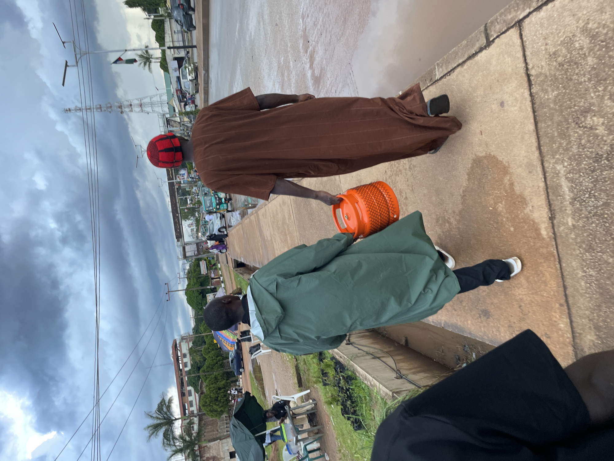 In Jos, Nigeria, two individuals walk along a concrete sidewalk adjacent to a street. The person on the left, facing away from the camera, wears a long green raincoat and dark pants, with white sneakers. The person on the right, also facing away, is dressed in a brown, striped garment and a red and black checkered cap, wearing black sandals. This individual carries a bright orange, cylindrical object, possibly a gas cylinder, with a netted texture. The environment appears overcast, with a cloudy sky. The street is wet, with puddles indicating recent or ongoing rainfall. In the background, there are various vehicles, buildings, palm trees, and a large telecommunications tower, suggesting an urban setting. Some smaller plants in black plastic bags are visible on the left side of the sidewalk.
