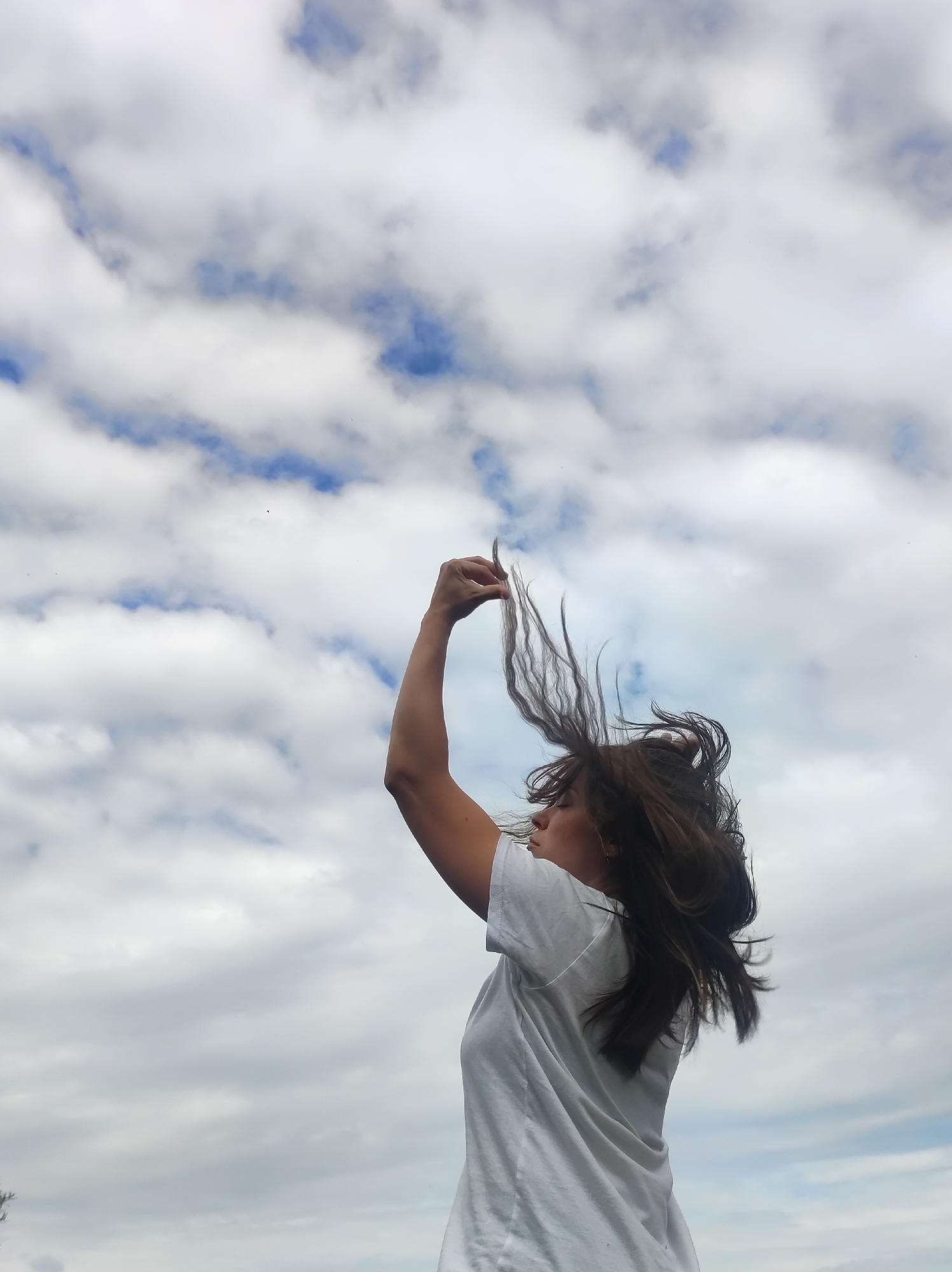 A woman is depicted from the mid-torso up, viewed from her right side against a bright sky with scattered clouds. Her long brown hair is dramatically lifted and fanned out upwards and to the viewer's left, appearing to be in motion. She wears a plain white short-sleeved t-shirt. Her right arm is raised, with her hand positioned above her head, seemingly interacting with her hair. Her face is turned to the left in profile, with her eyes appearing closed. The background consists entirely of a bright blue sky partially covered by white cumulus clouds. The location is the city of Unknown, Unknown.