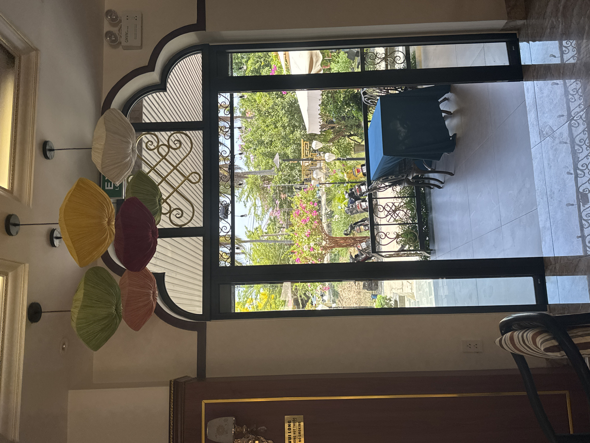 A view through an arched doorway shows a table on a terrace, an external garden, and objects hanging on the interior wall, likely in Đà Nẵng, Vietnam. The doorway has a decorative wrought iron design. Outside, a wrought iron table and chairs are partially covered with a blue cloth. The garden beyond contains tropical plants, some with pink flowers, and possible street lighting. Inside, hanging decorations of various colors are affixed to the wall to the left of the doorway. The lower part of the doorway is framed by a dark wood panel. Part of a chair is visible on the right side. Light patterns illuminate the tiled floor of the terrace.