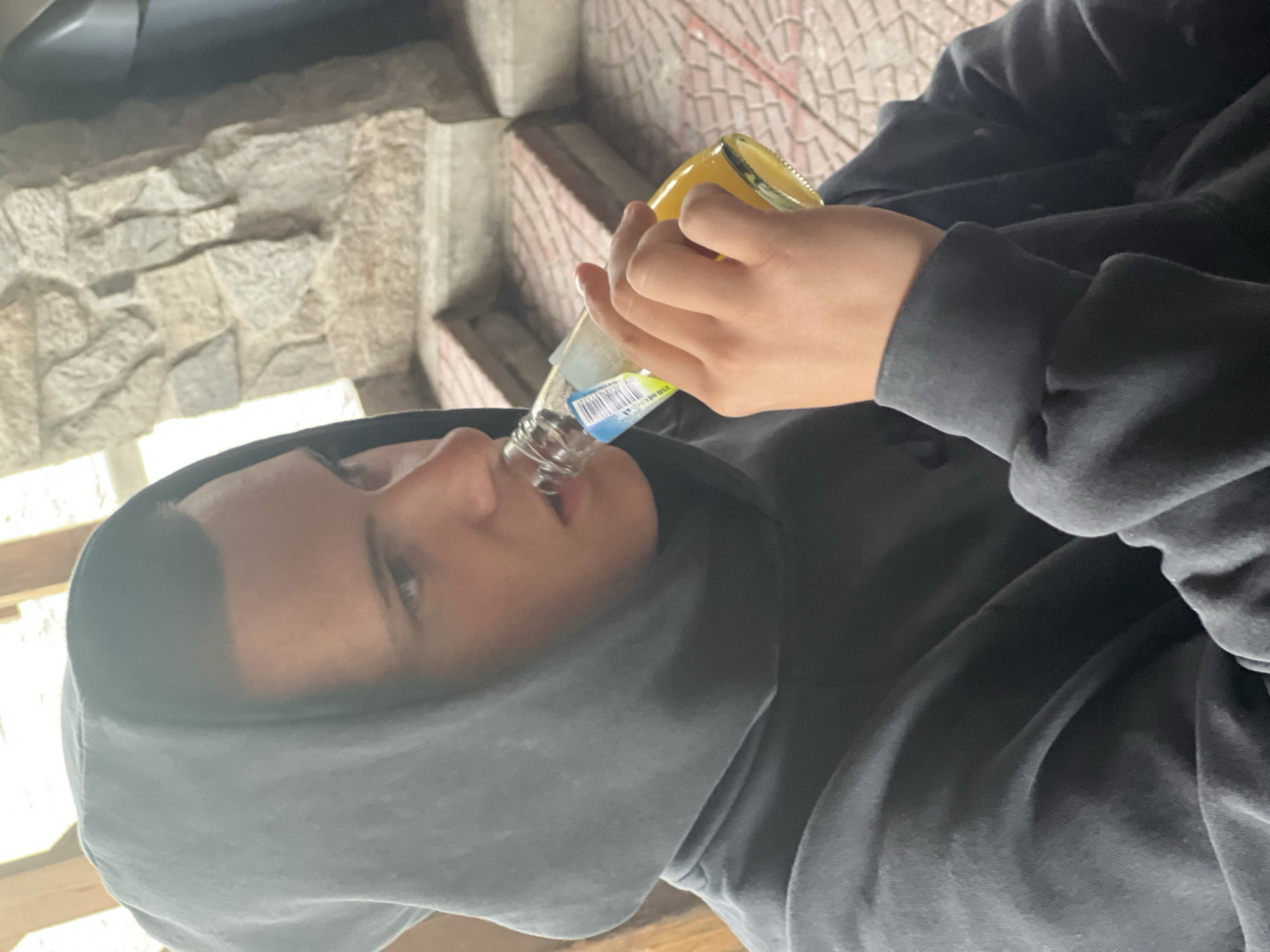 A person wearing a gray hooded sweatshirt is drinking from a glass bottle with a yellow liquid inside. The bottle is held in both hands. The background includes a wall constructed of stone and a tiled surface. The location is unspecified, but the image description indicates the city of Asenovgrad, Bulgaria.