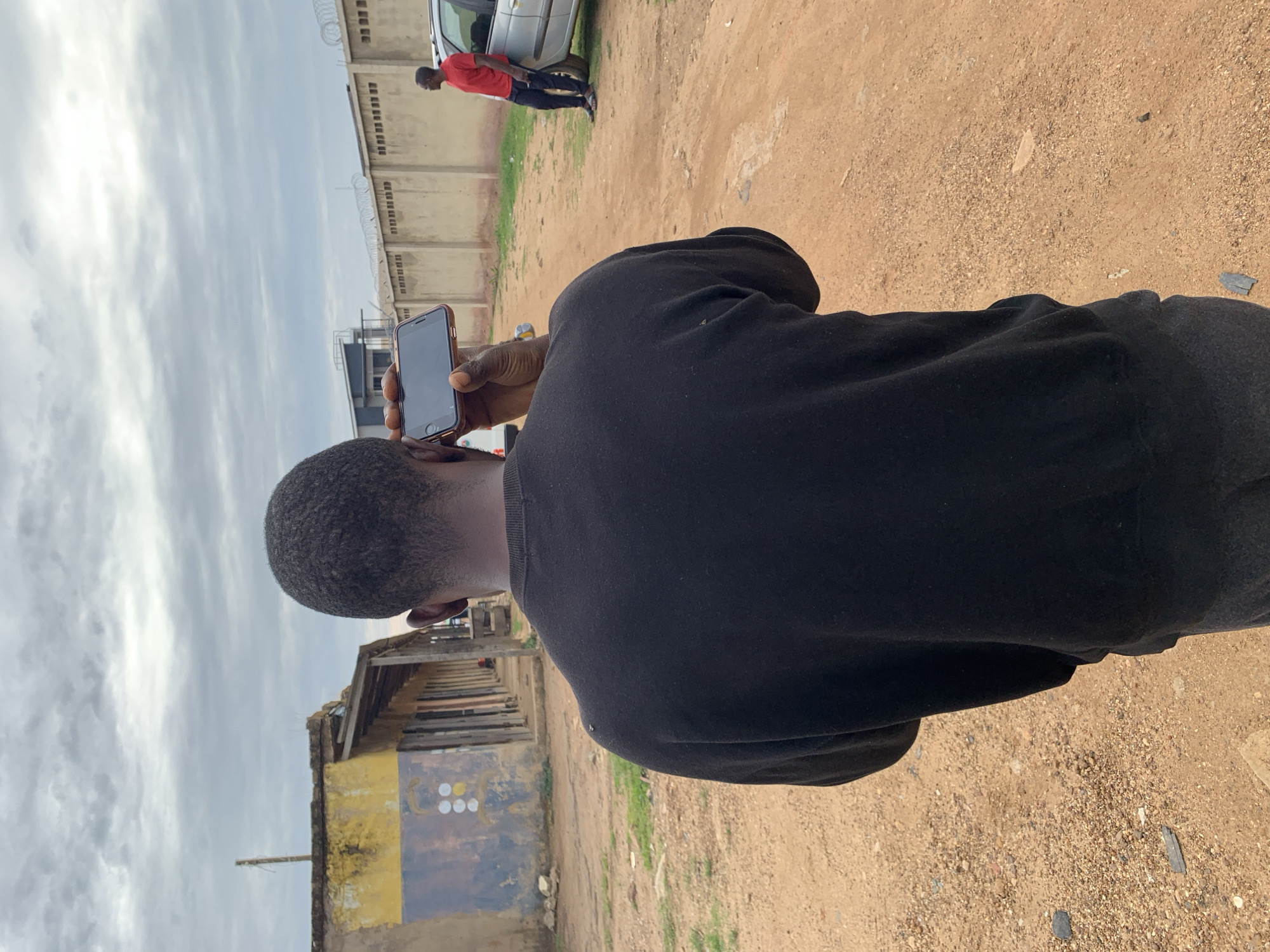The image depicts an outdoor scene in Rafin Par, Nigeria, under an overcast sky. The primary subject is a dark-skinned individual viewed from behind, wearing a black long-sleeved sweatshirt and dark trousers. This individual holds a black smartphone horizontally in their right hand, with the screen facing away, suggesting they are either recording or photographing.
The foreground is a reddish-brown dirt and gravel path with scattered small rocks and sparse patches of green vegetation. To the left, a series of single-story, dilapidated structures are visible. These buildings exhibit faded paint in yellow, blue, and brown tones, with visible wooden framing and doorways. A utility pole or similar wooden post stands vertically near the leftmost structure.
In the mid-ground on the right, another dark-skinned individual is seen wearing a red t-shirt, dark trousers, and sandals. This person leans against the front portion of a silver sport utility vehicle (SUV) parked on the dirt path.
Behind the person on the right, a tall, light-colored concrete perimeter wall is present, topped with multiple strands of razor wire or barbed wire. Beyond this wall, a modern building with a dark roof and lighter-colored walls is partially visible against the cloudy horizon.