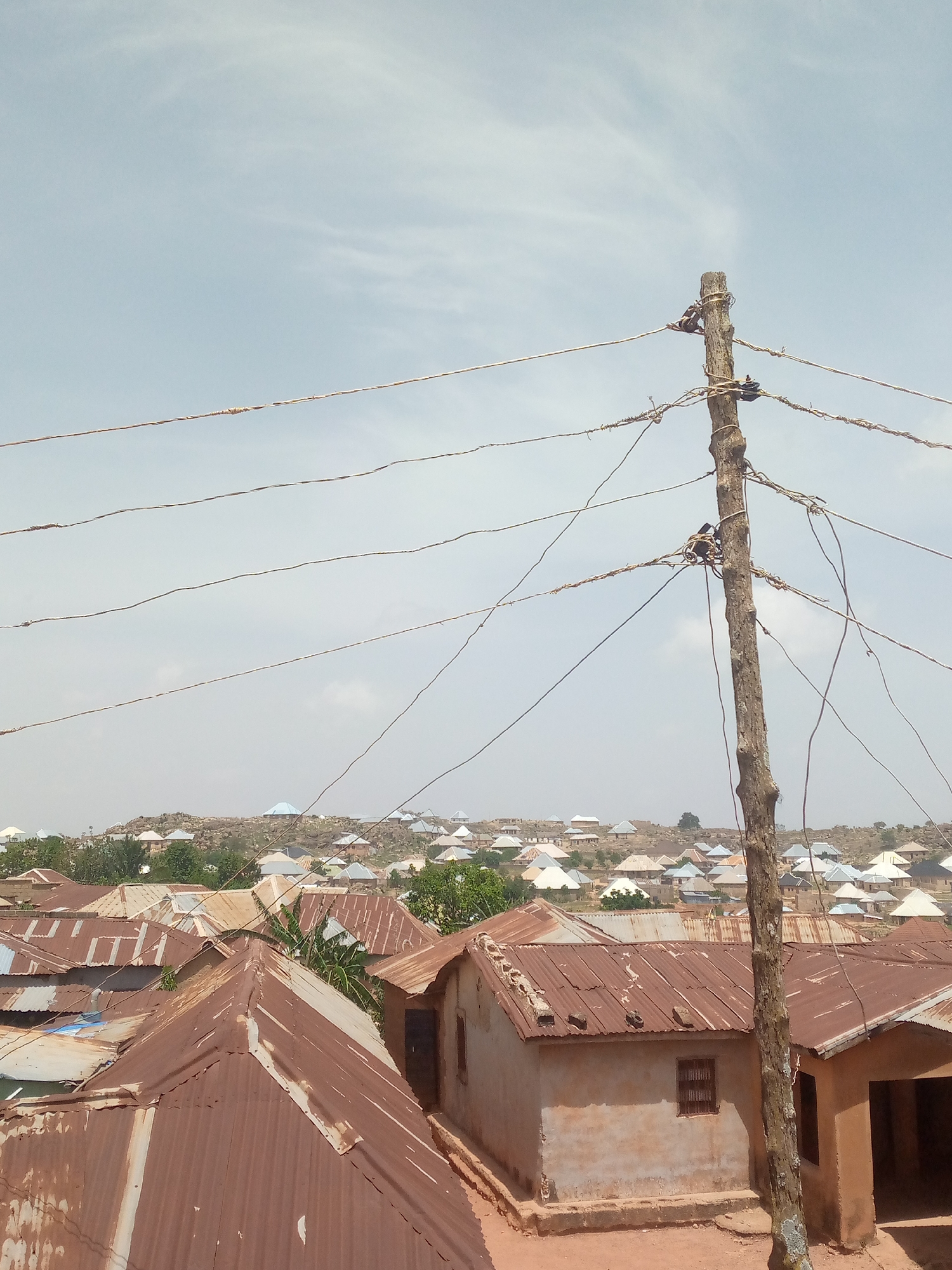 The image depicts a scene in Bukuru, Nigeria. In the foreground, corrugated metal roofs of buildings dominate, some with rust. A weathered wooden utility pole stands prominently, with multiple wires haphazardly strung across it. Beyond the foreground buildings, a hillside is covered with more structures, varying in color and roof style. The sky is overcast. No people are visible.
