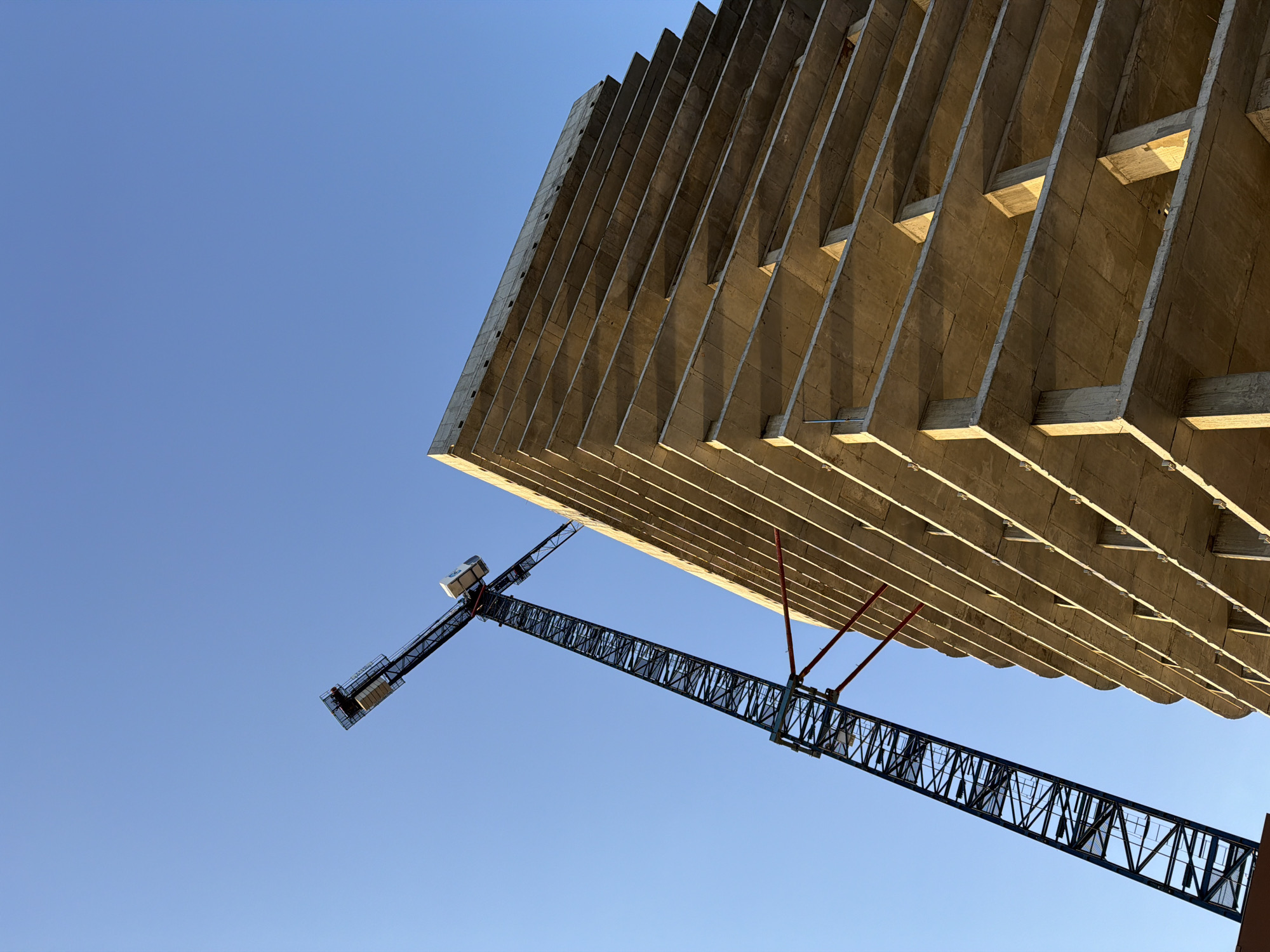The image captures a low-angle view of a construction site in Kazan, Russia. A concrete building, still under construction, dominates the upper portion of the frame. The building features a layered design with protruding concrete supports. A dark-colored construction crane stands alongside the building, its arm extending toward the left side of the image. The sky is a clear, bright blue.