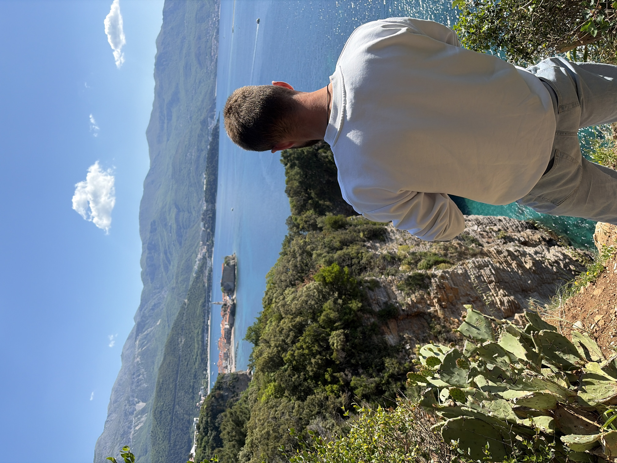 The image shows a person, presumably male, seen from the back, leaning towards a cliff edge overlooking the Adriatic Sea. The person is wearing a white sweatshirt and light-colored jeans. The scene is located in Budva, Montenegro, as evidenced by the view of the coastline, surrounding mountains, and buildings in the distance. Below the cliff, there is a lush green area with trees and cactuses. The sea is calm, with some boats visible. The sky is mostly clear with a few scattered clouds. The overall lighting indicates a sunny day.