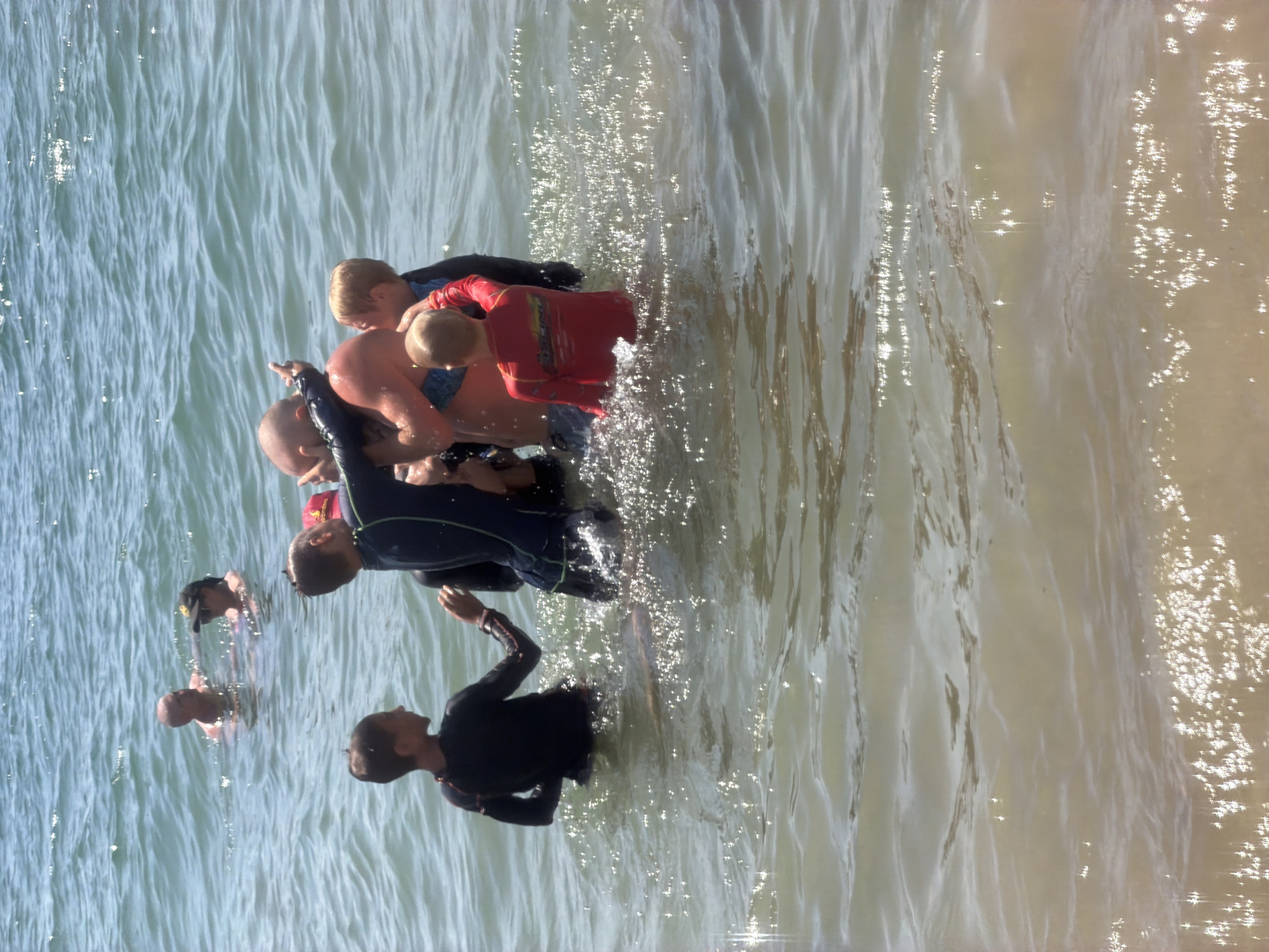This is a vertically oriented, top-down, wide shot of a group of people in the ocean, likely during a surfing or swimming lesson. The water is shallow and appears to be a beach in Nha Trang, Vietnam. The sun is shining, as evidenced by the sparkles on the water, suggesting it is daytime, likely late morning or early afternoon.
The subjects are primarily children and at least one adult. Several children are wearing wetsuits, some red and some blue. One child is in a red wetsuit, and another is in a blue one. There's a man with a bald head in the water. Another child, seen from the back, is wearing a black wetsuit. The main activity seems to be instruction in the water, with the adult possibly guiding or teaching the children. Some of the children are clustered together, while others are more spread out. The water is clear and appears calm with gentle ripples. The overall atmosphere seems positive and active, with the subjects engaged in a water sport. There are no visible objects other than the people and the water. No text is visible in the image.