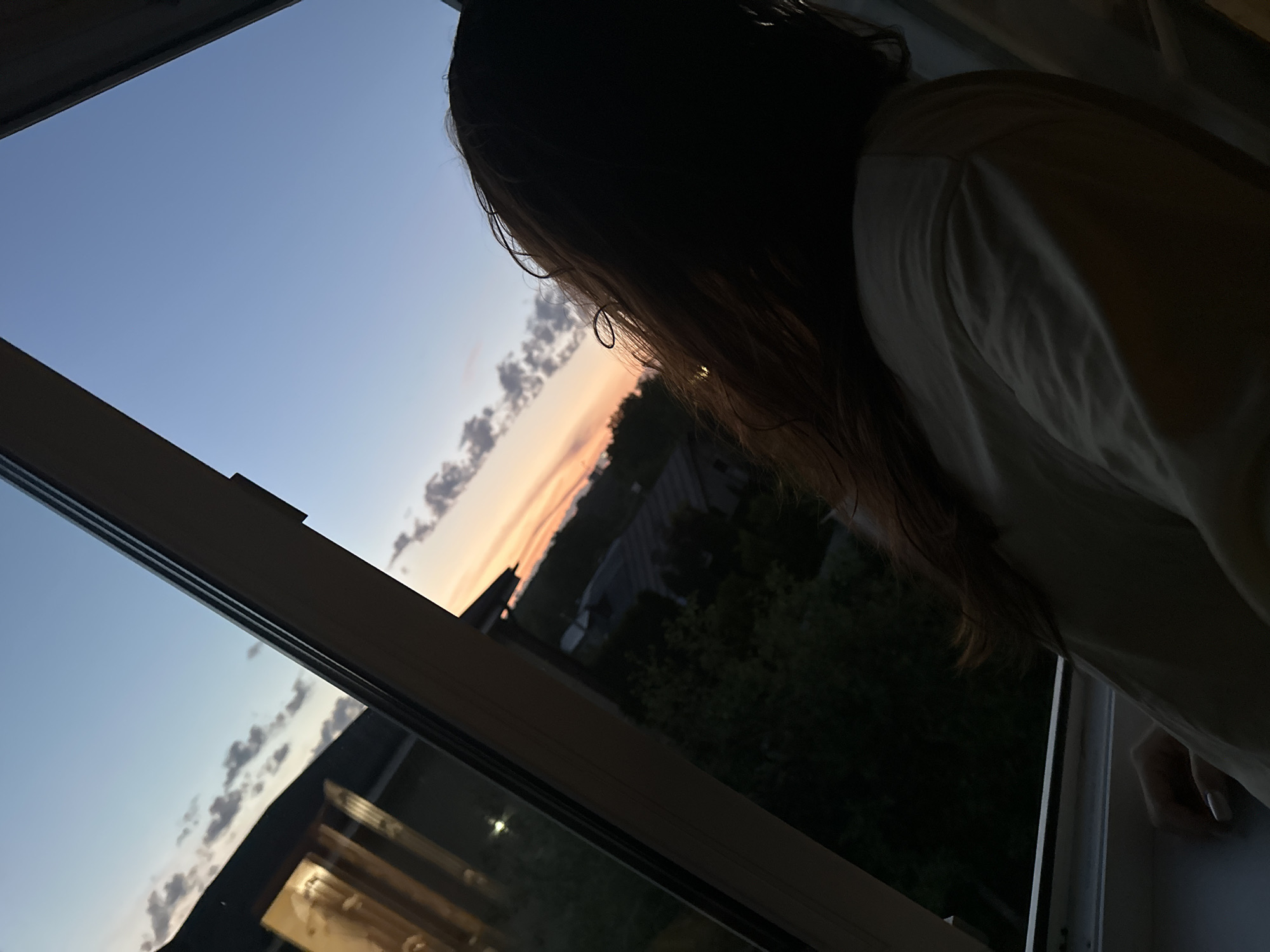 A young woman with dark hair, seen from behind, gazes out of a window at a sunset over a rural landscape, possibly on the outskirts of Nizhny Novgorod, Russia. The sky displays a gradient of blue to orange, with scattered clouds. Outside, trees and structures are visible in the fading light. The woman is wearing a light-colored shirt, and her left hand rests on the windowsill, with painted fingernails. The scene is viewed from inside, with the window frame prominent in the foreground.