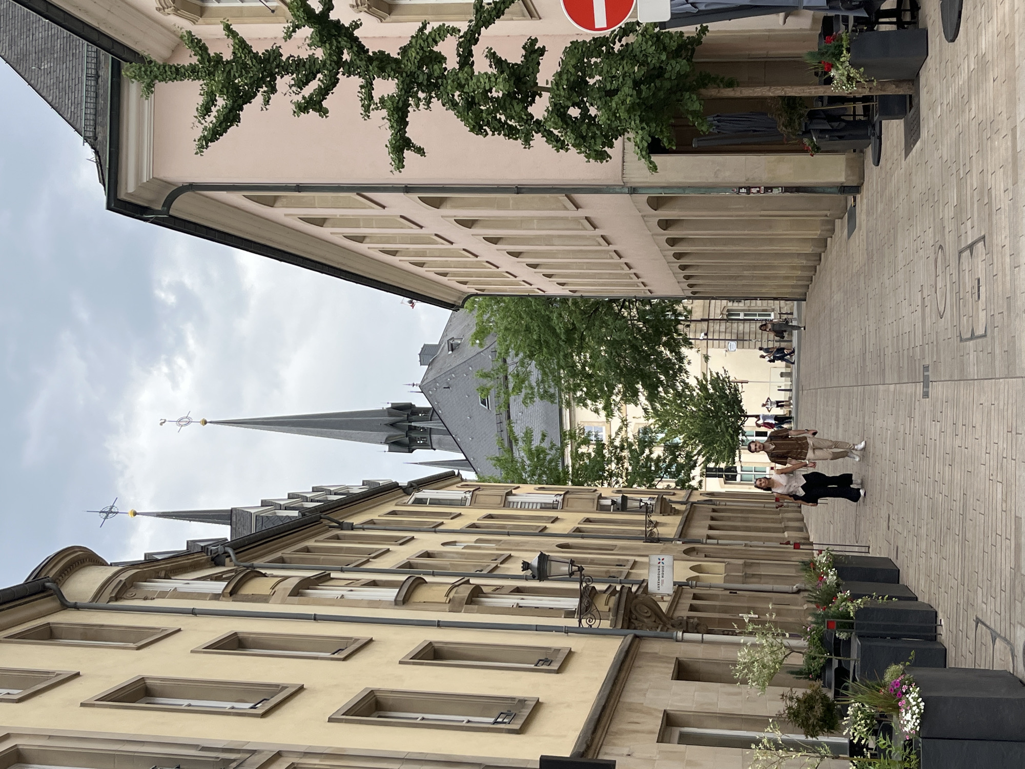 An outdoor, eye-level, medium shot captures a narrow street in an urban environment. The street is paved with light-colored bricks and lined with buildings on both sides. The building on the left is tall and has a pale yellow facade with multiple windows. The building on the right is shorter and painted a light pink. Two spires of a church or cathedral rise above the buildings in the background, with a cloudy sky overhead.
In the foreground, a couple walks hand-in-hand down the street, facing away from the camera. The woman wears a white vest and black pants, while the man is dressed in a brown shirt and light-colored pants. Planters with greenery and flowers are placed along the left side of the street. Further down the street, several people are walking, and outdoor seating areas with umbrellas are visible on the right. A red and white circular sign with a horizontal bar is affixed to a pole on the far right. The overall atmosphere appears calm and leisurely.