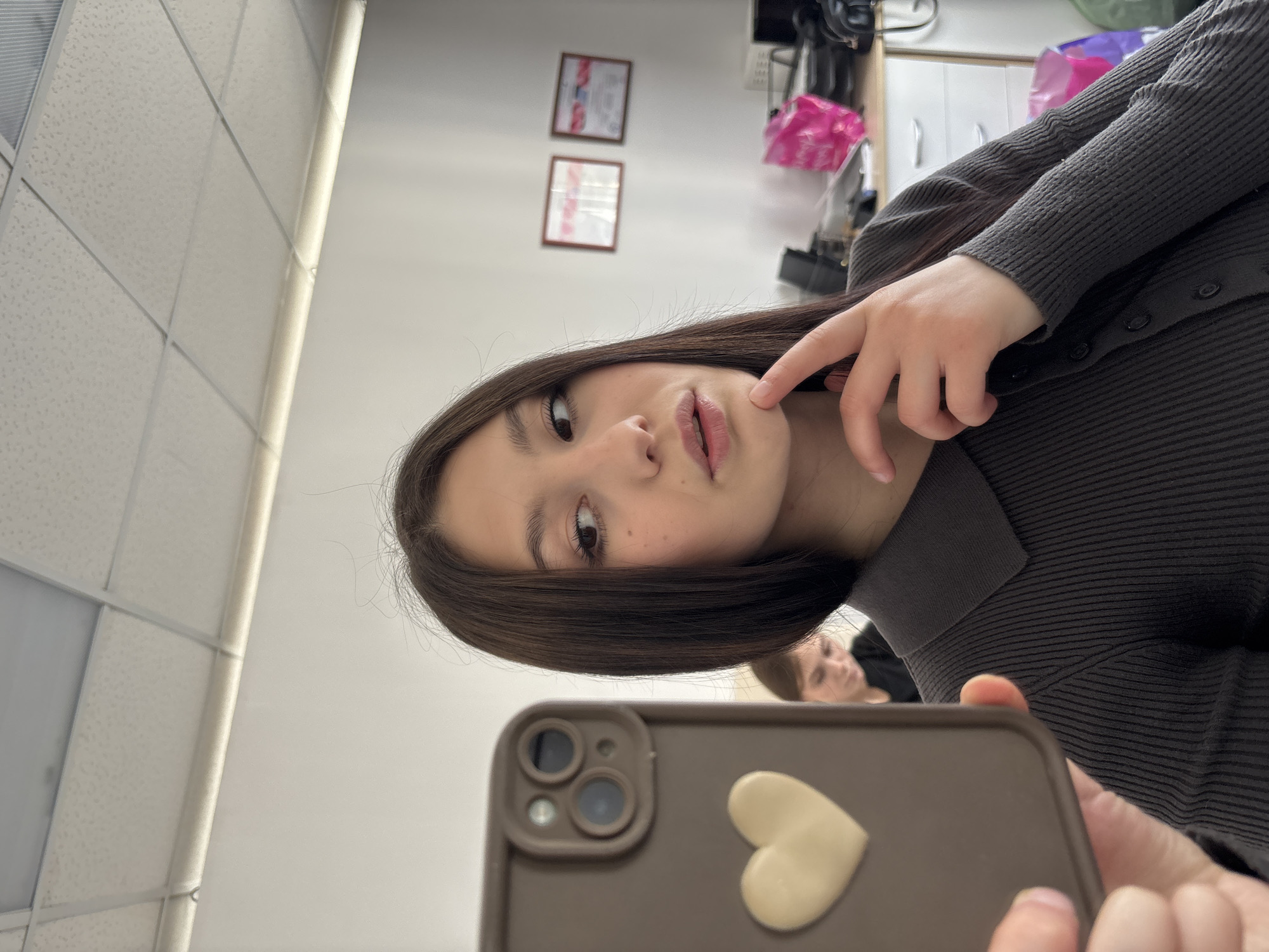 A young woman is taking a selfie indoors, holding a phone with a brown case and a heart-shaped sticker. She is touching her cheek with her finger. Her hair is short and dark brown. She is wearing a dark gray sweater with buttons. The background shows a white wall and ceiling with a grid pattern. In the background, behind her, another person can be seen. Two framed certificates are also visible on the wall. The location is Фоки, Russia.