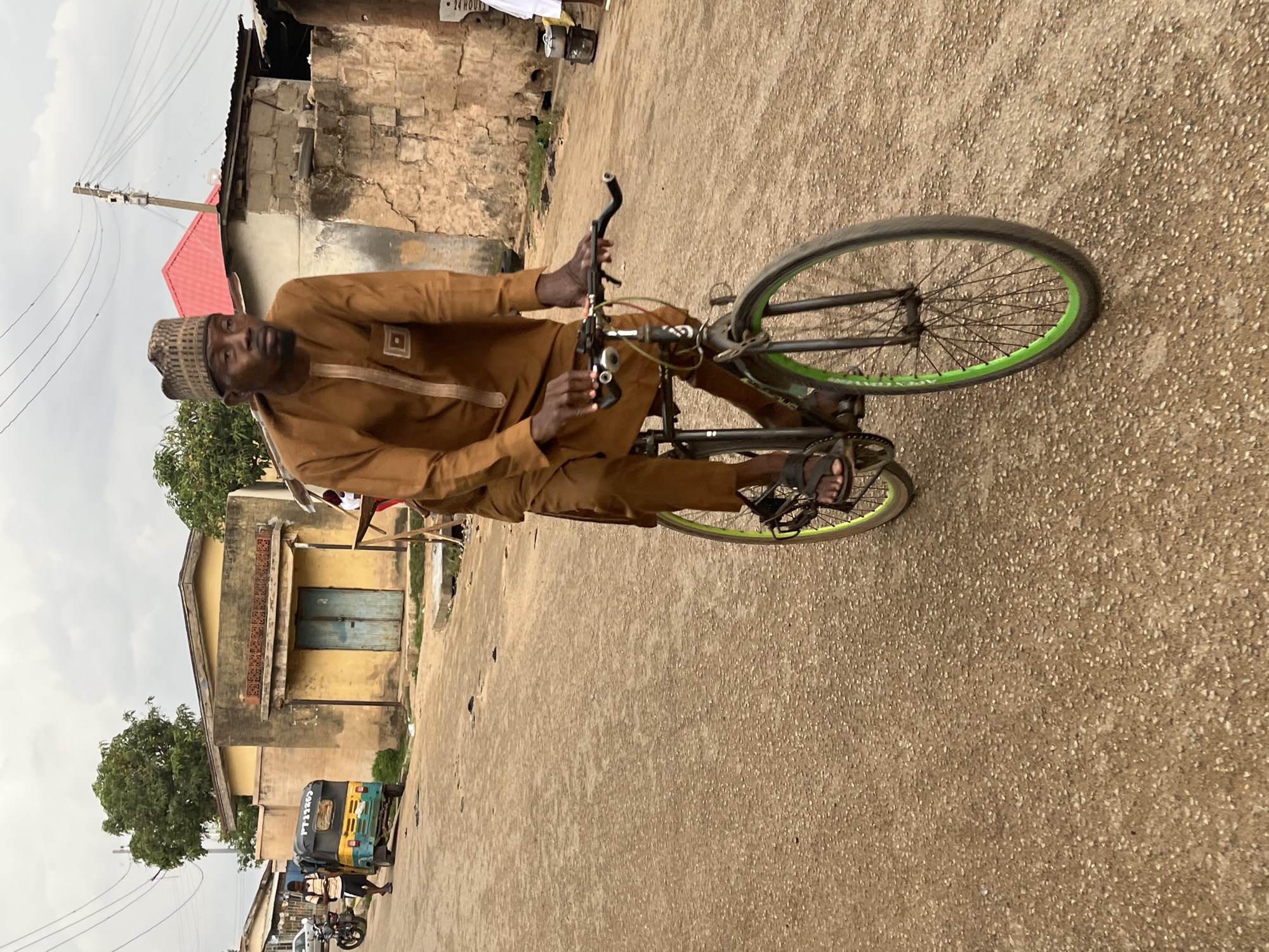The image depicts a man riding a bicycle on a paved road. The man is wearing a brown outfit and a patterned hat. The bicycle has green rims. The road is surrounded by buildings, including a yellow building with blue doors and a building with a red roof. In the background, a motorcycle and a three-wheeled vehicle, possibly a Bajaj, are parked. Based on the architectural style and the presence of the three-wheeled vehicle, the location is likely Jos, Nigeria.