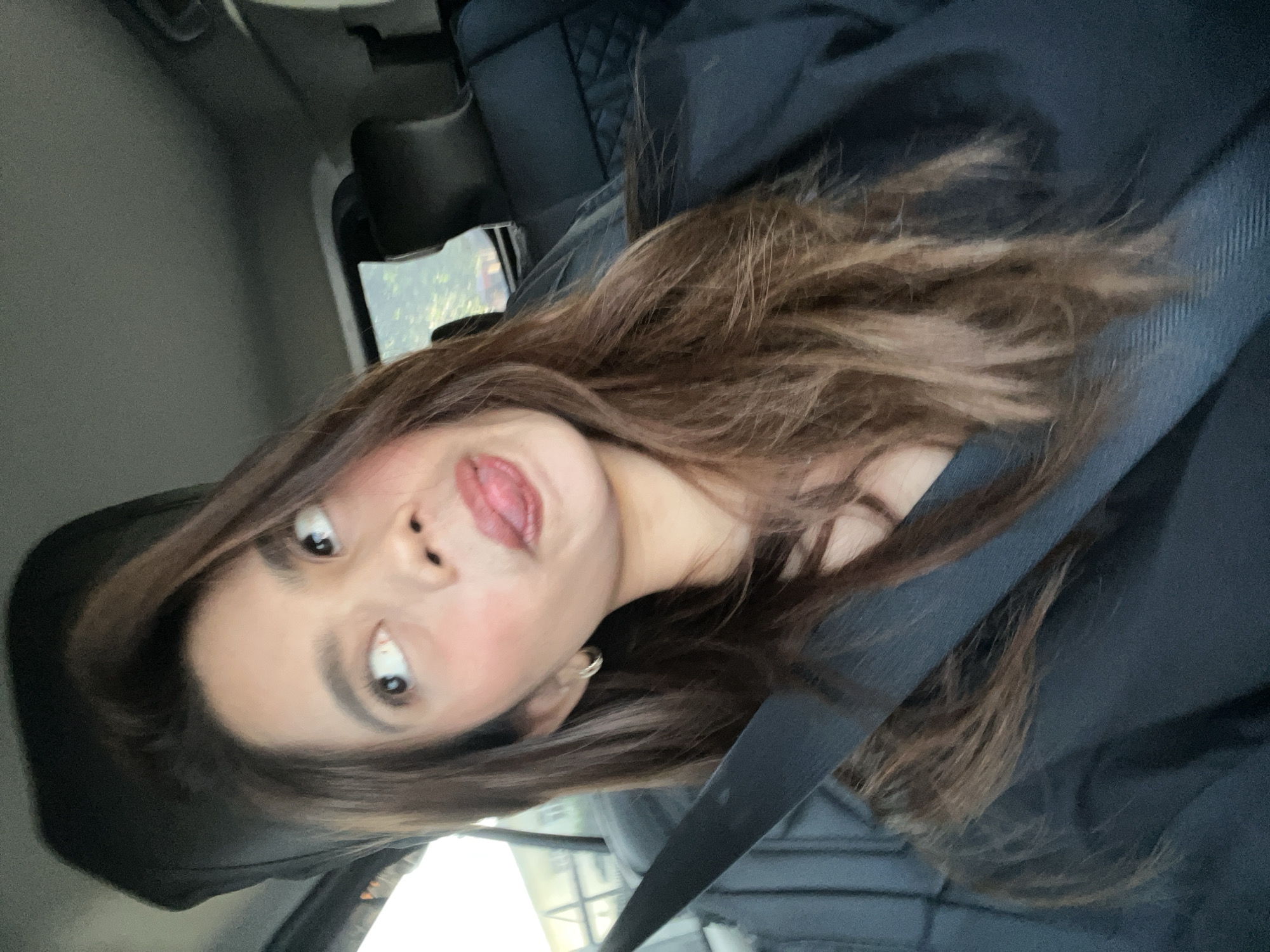 A female individual is seated in the front passenger seat of a vehicle, captured from the chest up. She has long, wavy, brown hair, brown eyes, and light skin, with visible blush on her cheeks. She is wearing a dark, possibly black, v-neck top and a black seatbelt across her left shoulder and chest. A small, silver-colored hoop earring is present on her left ear. Her mouth is open with her tongue extended, and her eyes are looking upwards and to her right. The interior of the vehicle is dark, with a dark grey or black ceiling visible at the top. Through the car windows, green foliage and trees are visible, indicating an outdoor, daytime setting. The rear seat upholstery is dark and features a quilted pattern. There are no specific visual identifiers within the image to confirm the location as Davao City, Philippines.